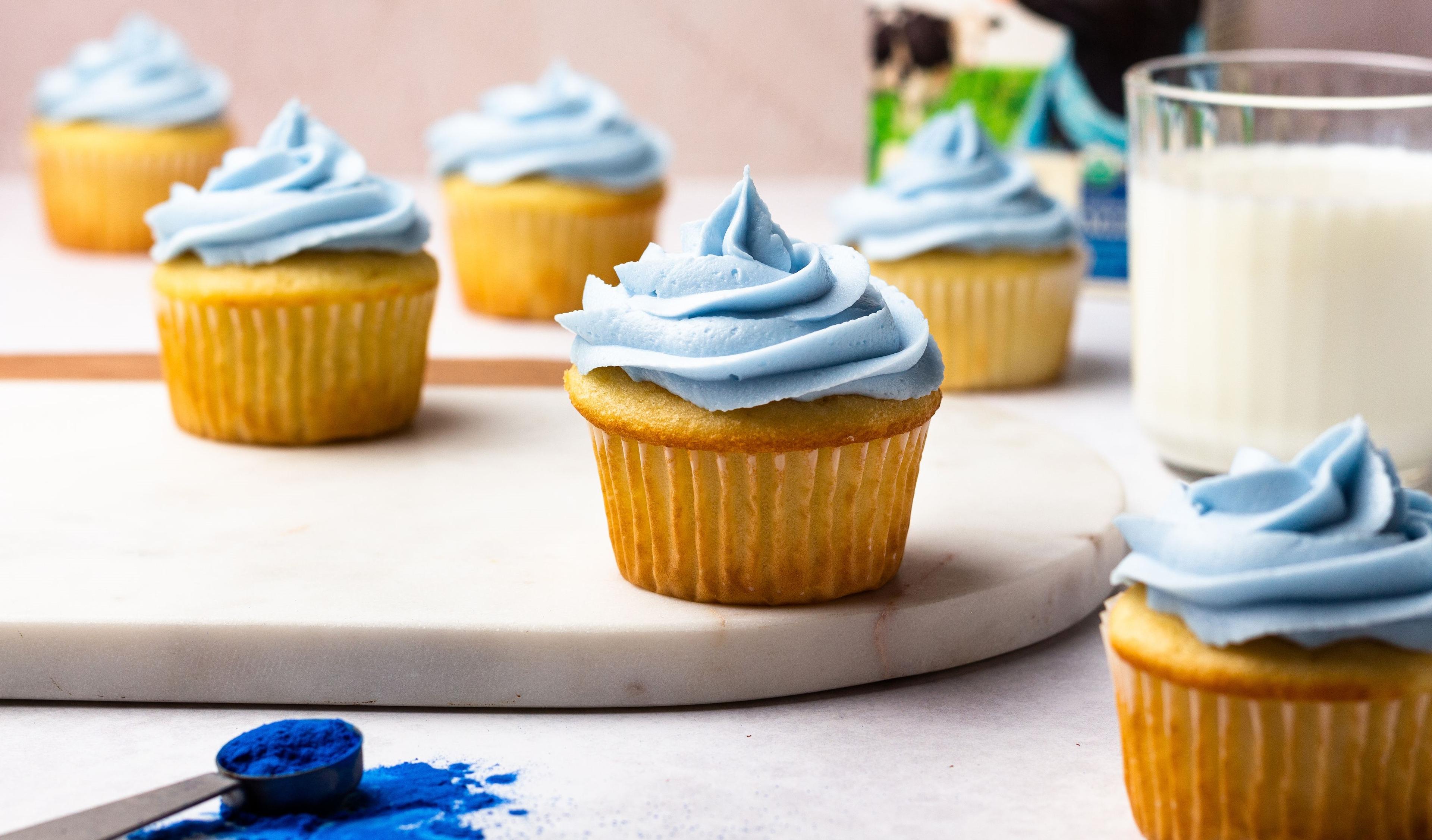 Blue spirulina is used for a natural frosting color on cupcakes placed next to a glass of milk.