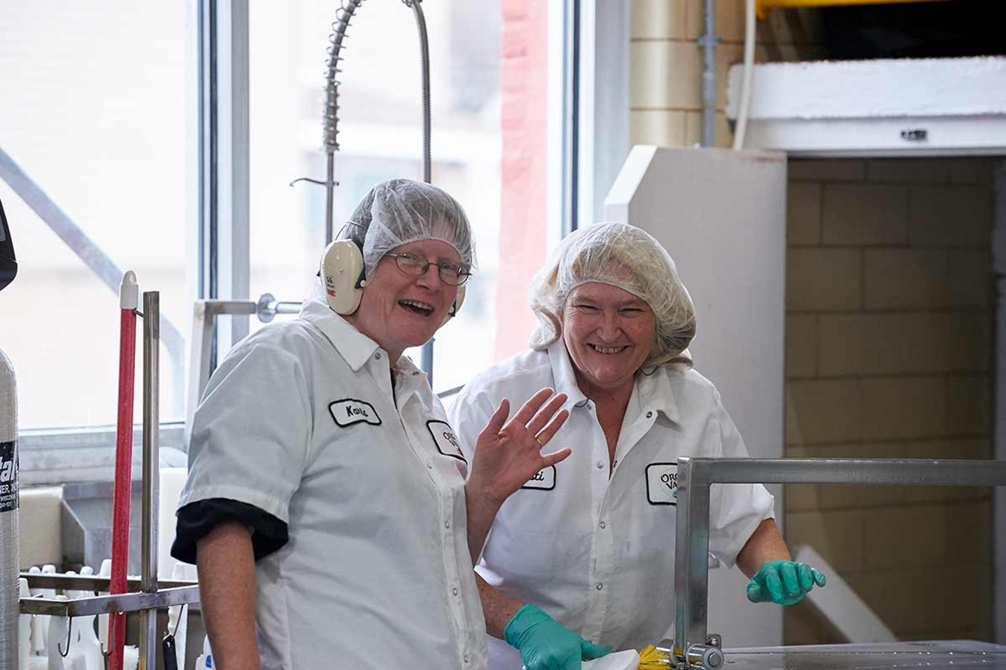 Two women in an Organic Valley production facility wearing white overcoats, hair nets and gloves smile and wave at the camera.