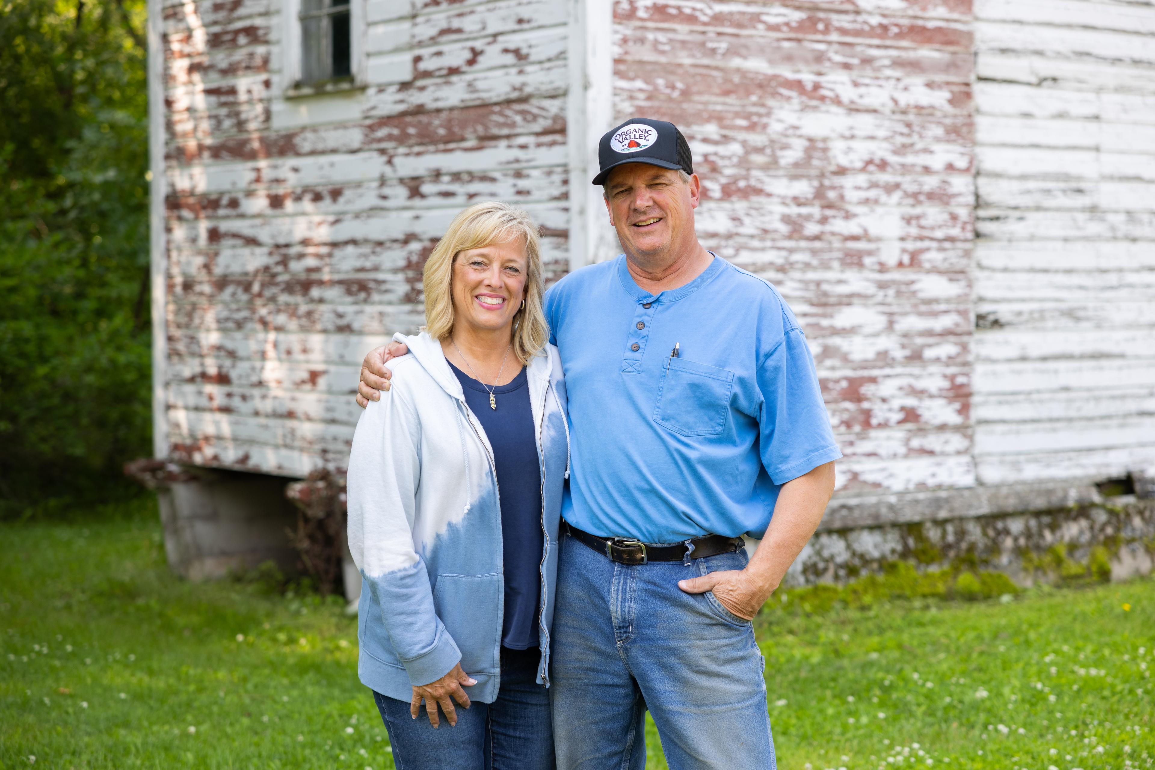 A husband and wife stand in front of a time-worn building on an organic farm. 