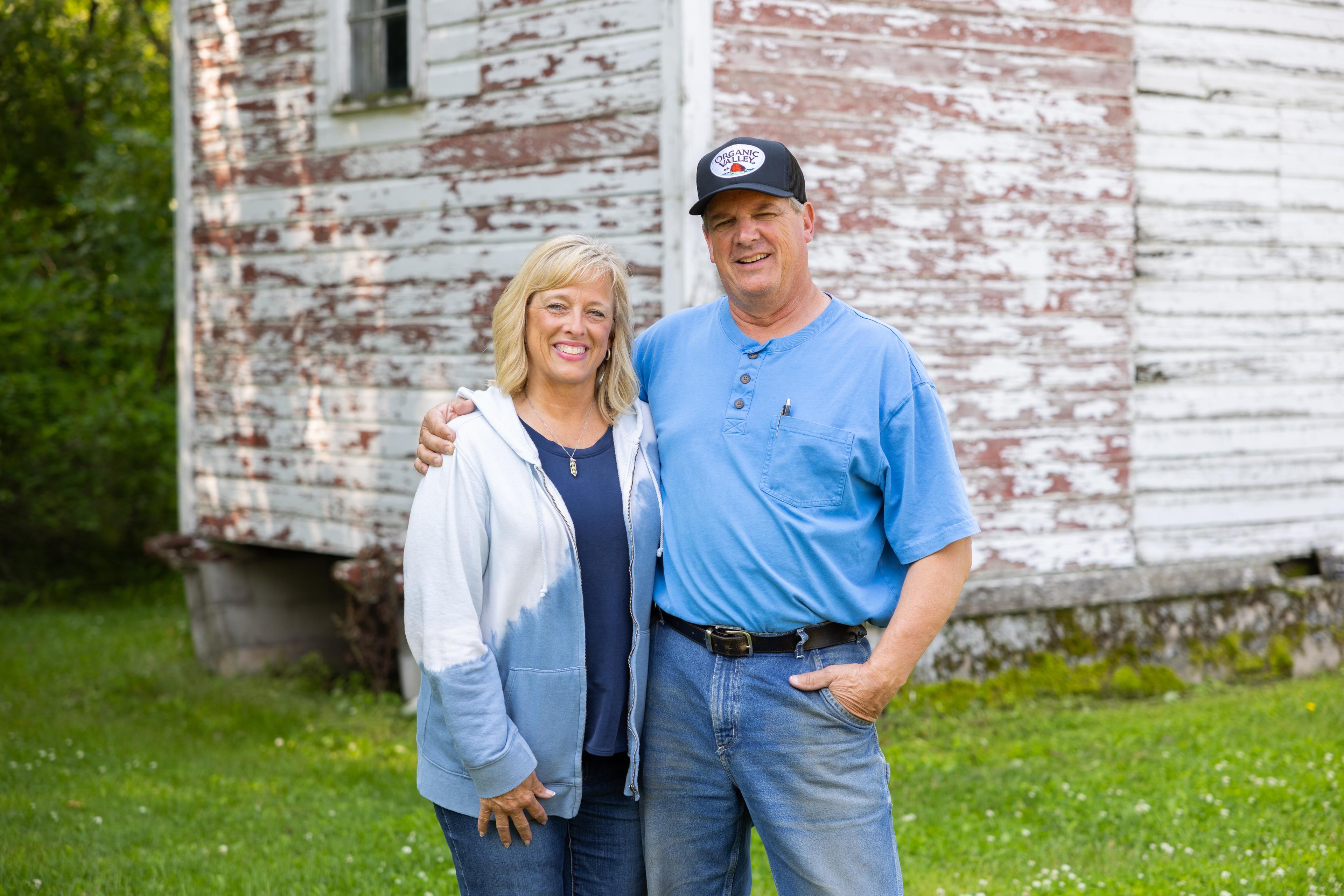A husband and wife stand in front of a time-worn building on an organic farm.