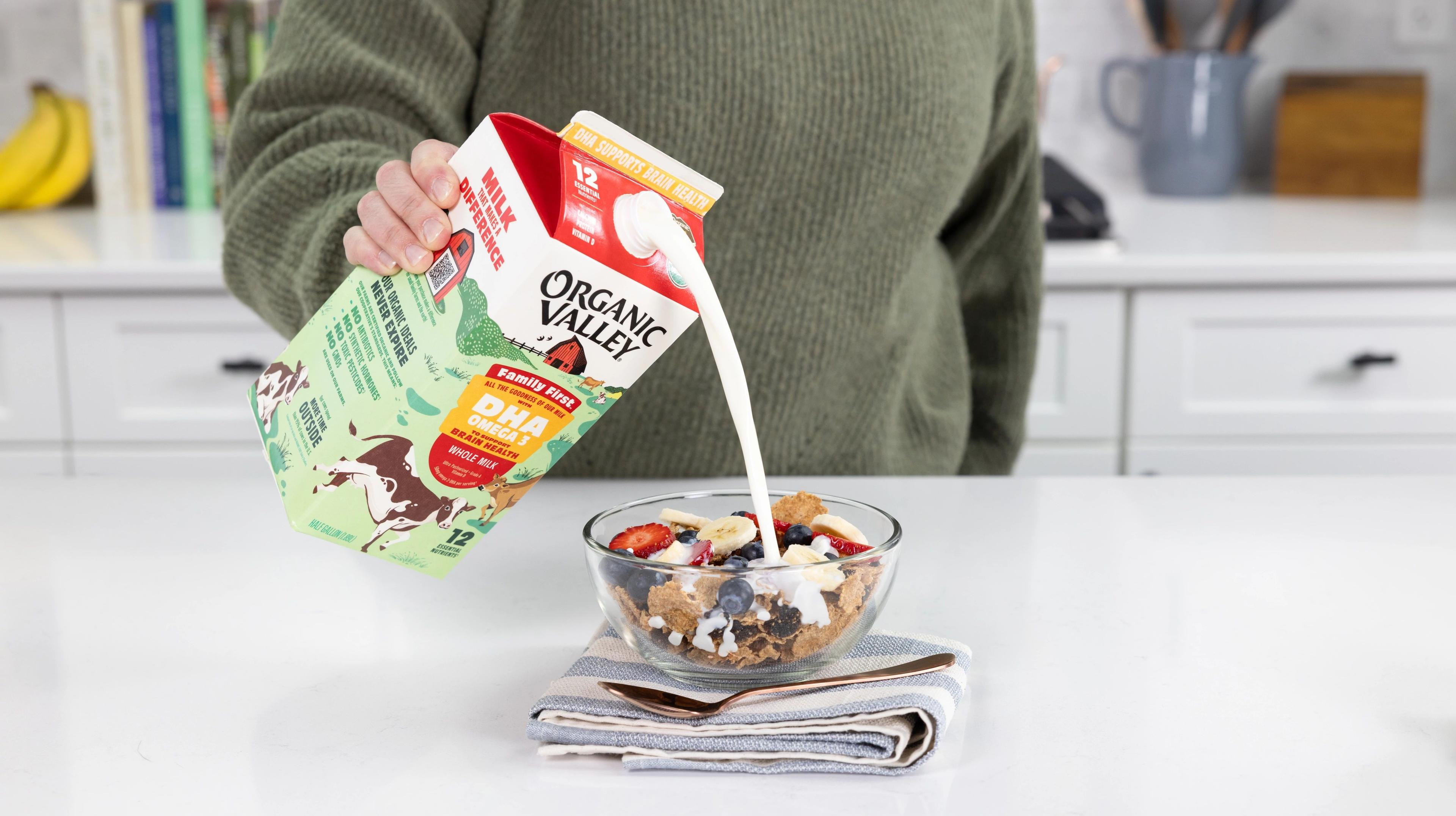 A person pours Family First organic milk into a bowl of cereal with strawberries, bananas and blueberries.