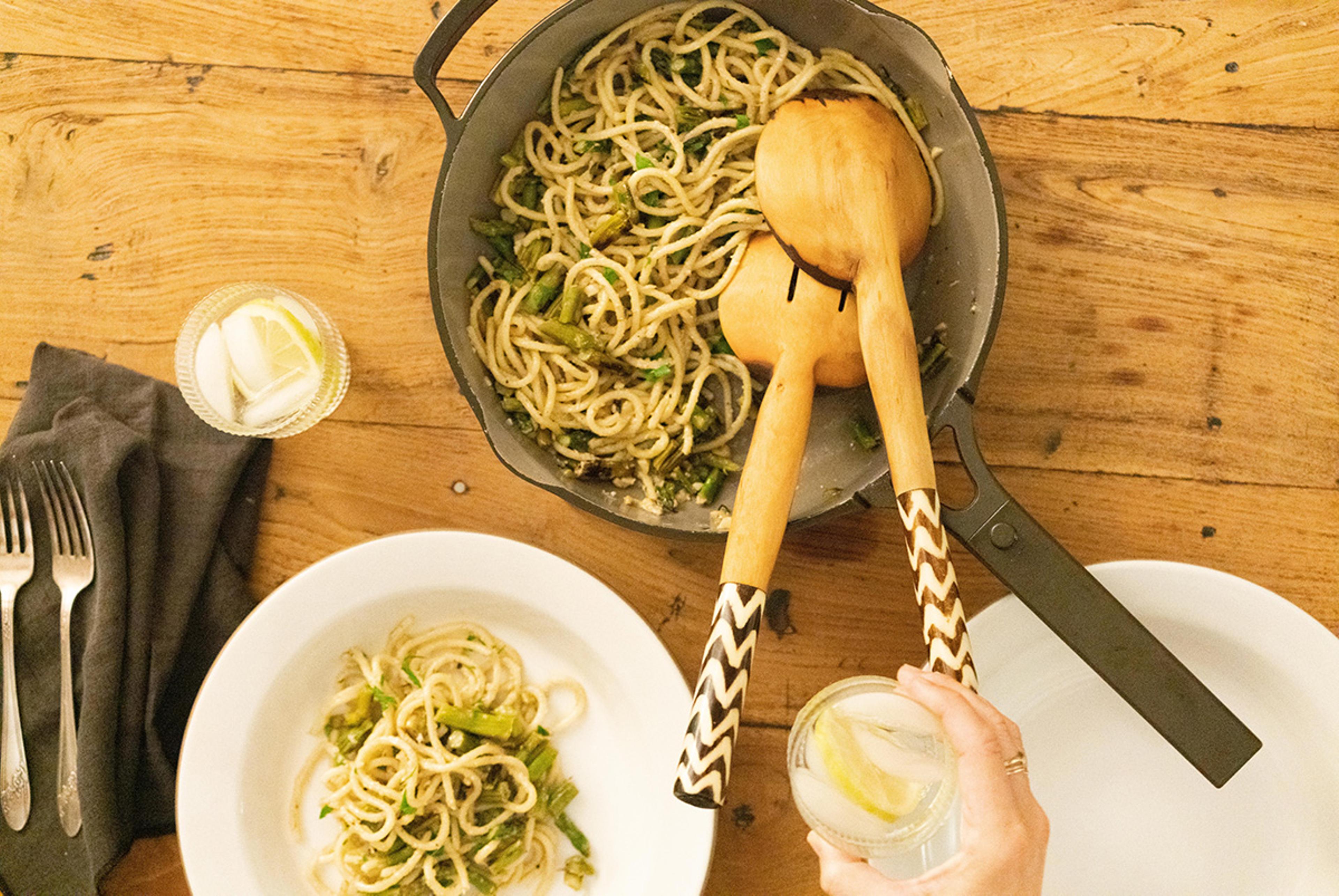 Overhead view of a healthy pasta dish with serving tongs and a hand holding a glass of water with lemon.