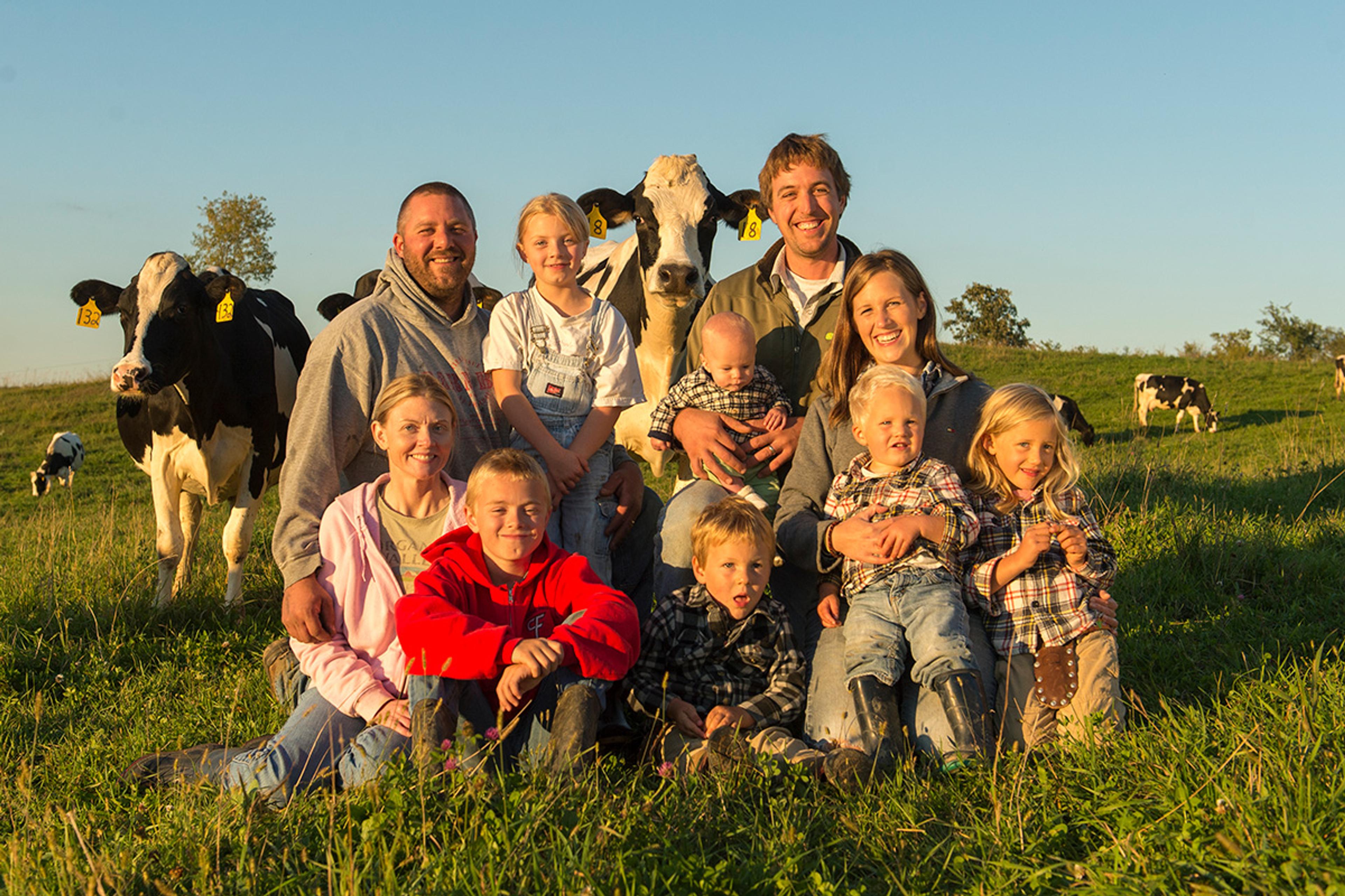 The O’Reilly family pose in a green pasture with cows and blue sky in the background.