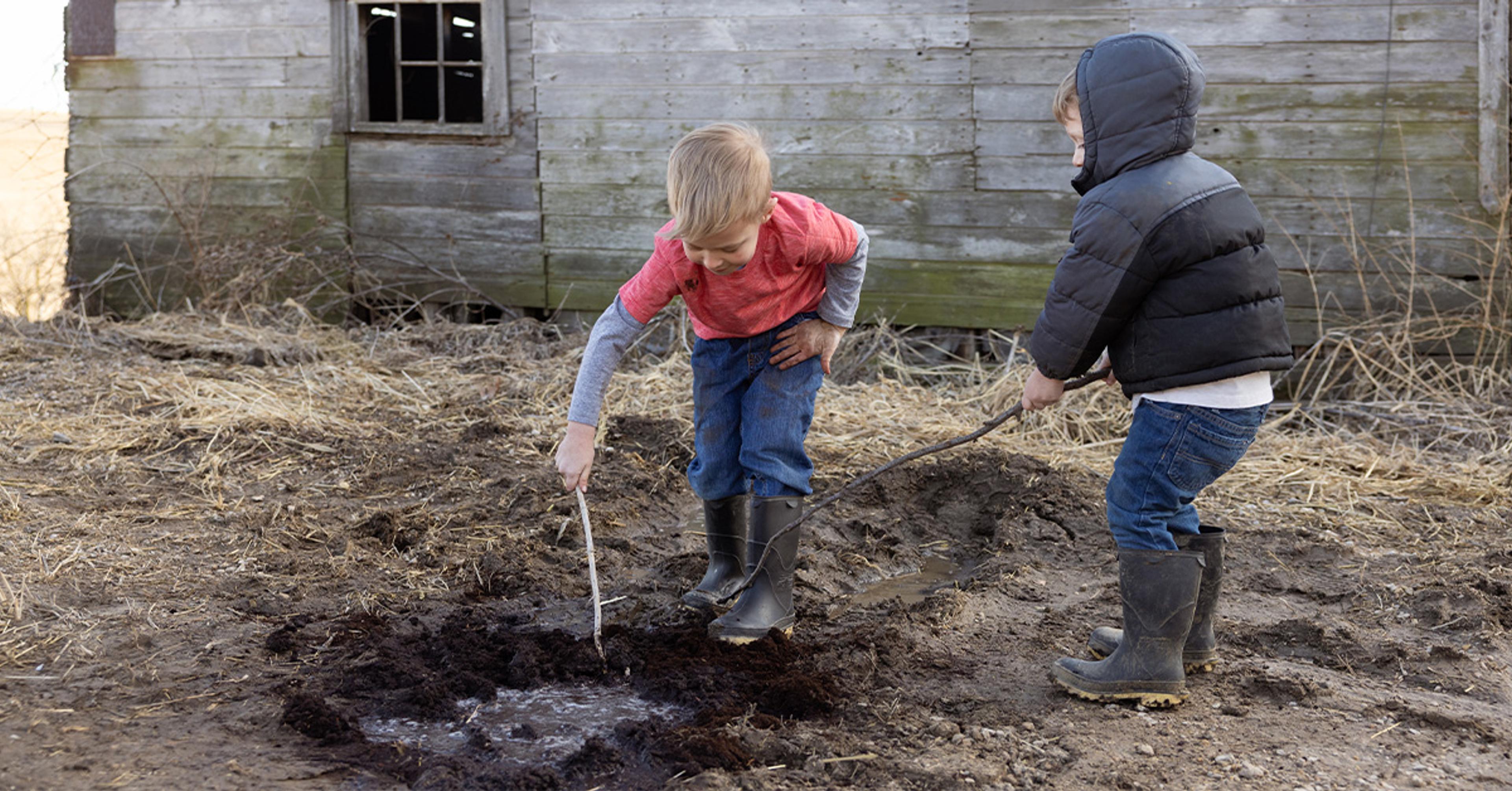 Two boys play with sticks in the mud in front of a barn. 