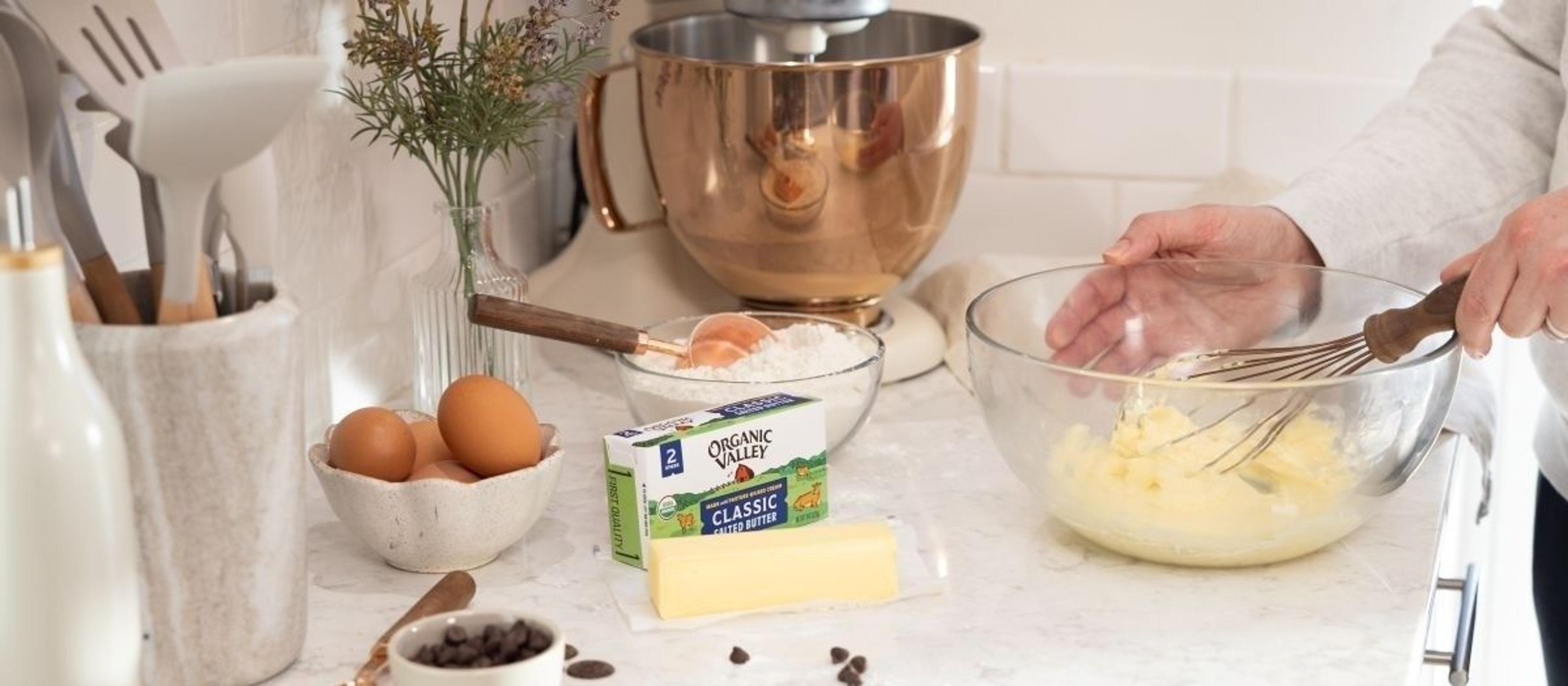 A woman blends butter in a bowl that’s on a counter; chocolate chips and baking ingredients are also on the counter.