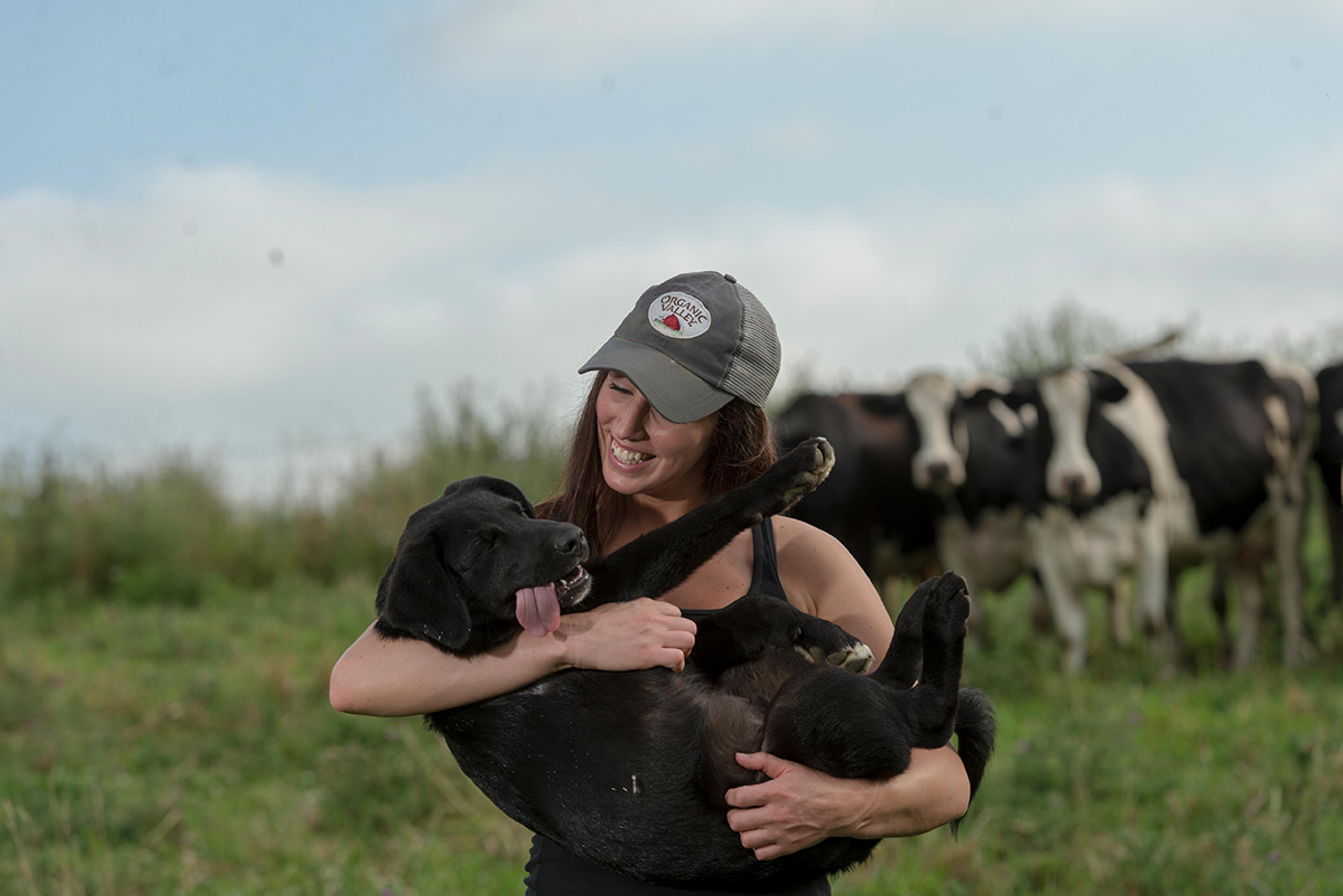 A young woman wearing an Organic Valley cap smiles down at a black puppy in her arms with its tongue hanging out.