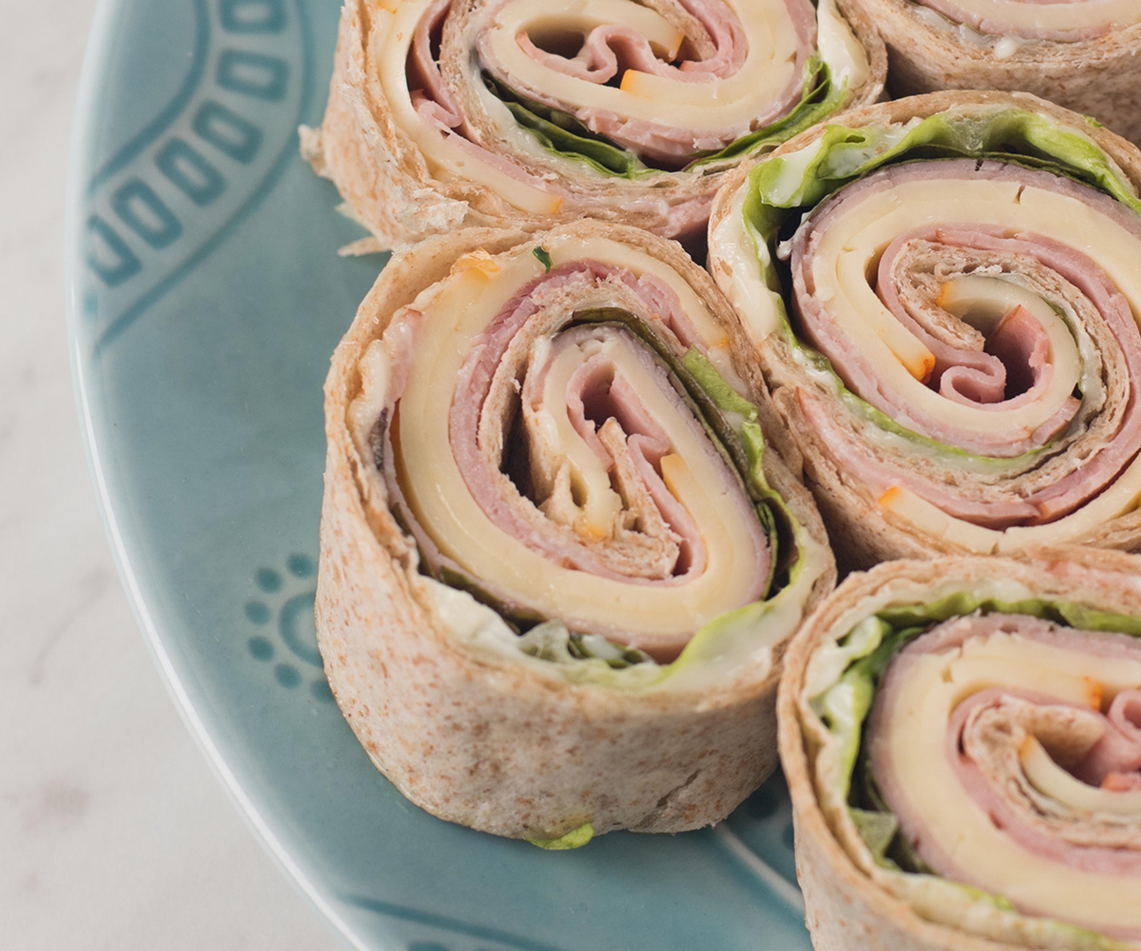 A wrap containing organic lunchmeat slices, sliced cheese and lettuce cut in slices and laid on a plate so they look like pinwheels.