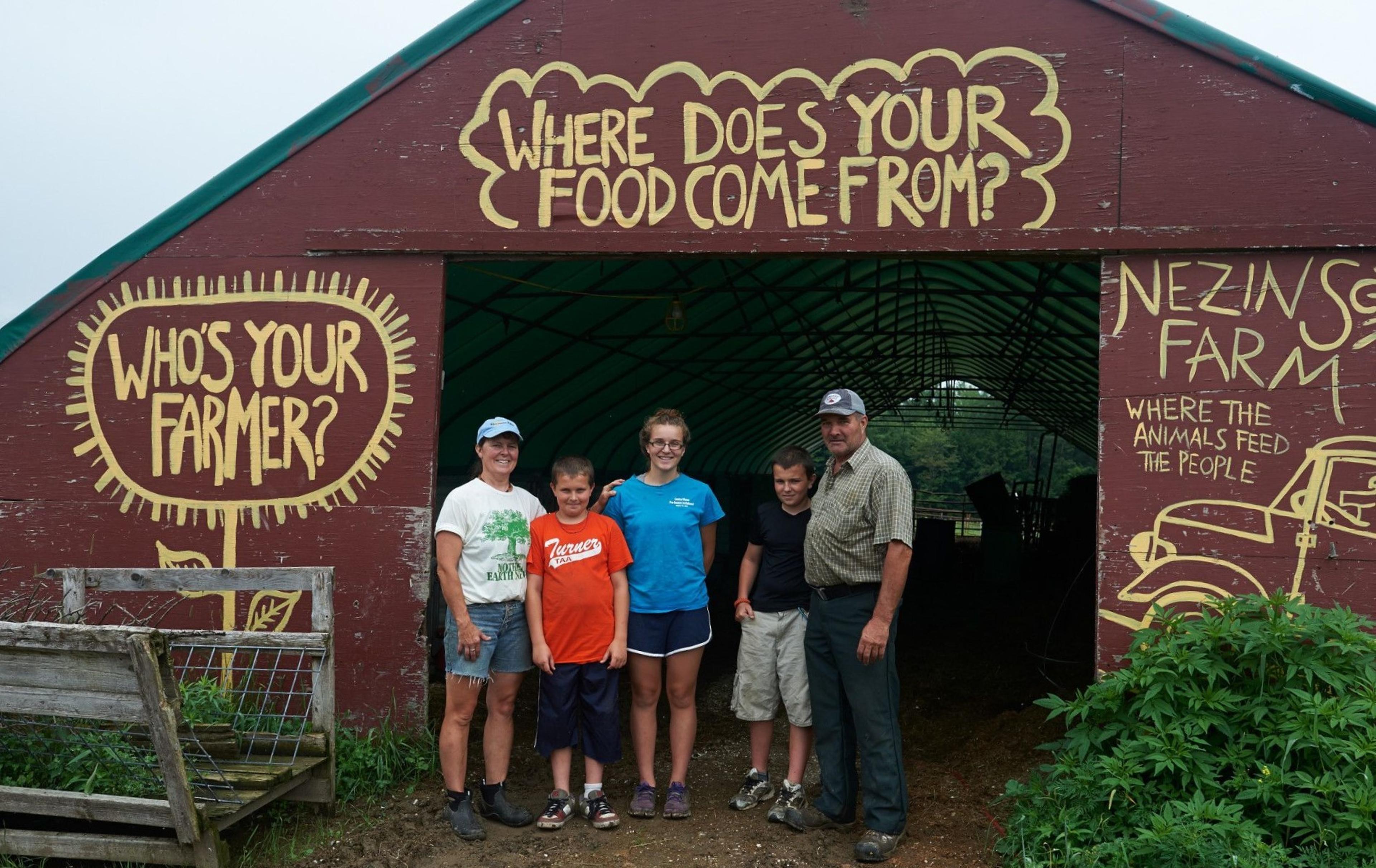 Five members of the Varney family in Maine stand in the doorway of a barn that has “who’s your farmer?” and “where does your food come from” painted on it.