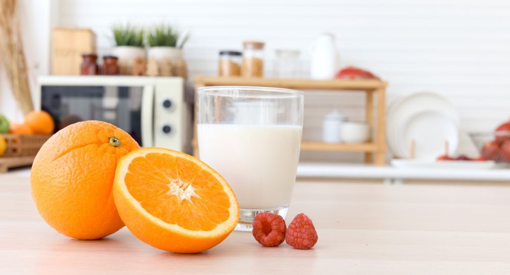 An orange, half and orange glass of milk, and raspberries on a kitchen counter.