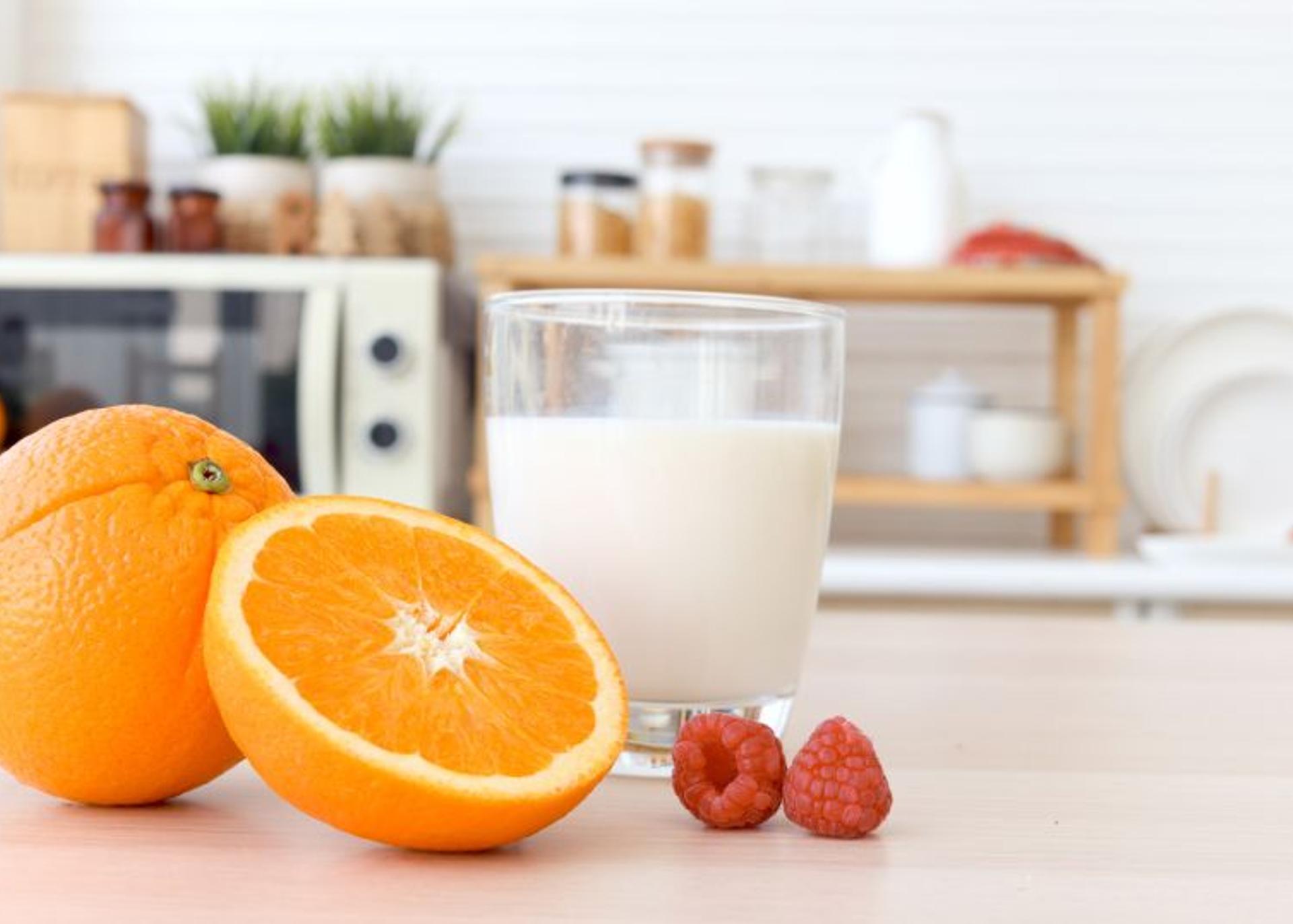 An orange, half and orange glass of milk, and raspberries on a kitchen counter.