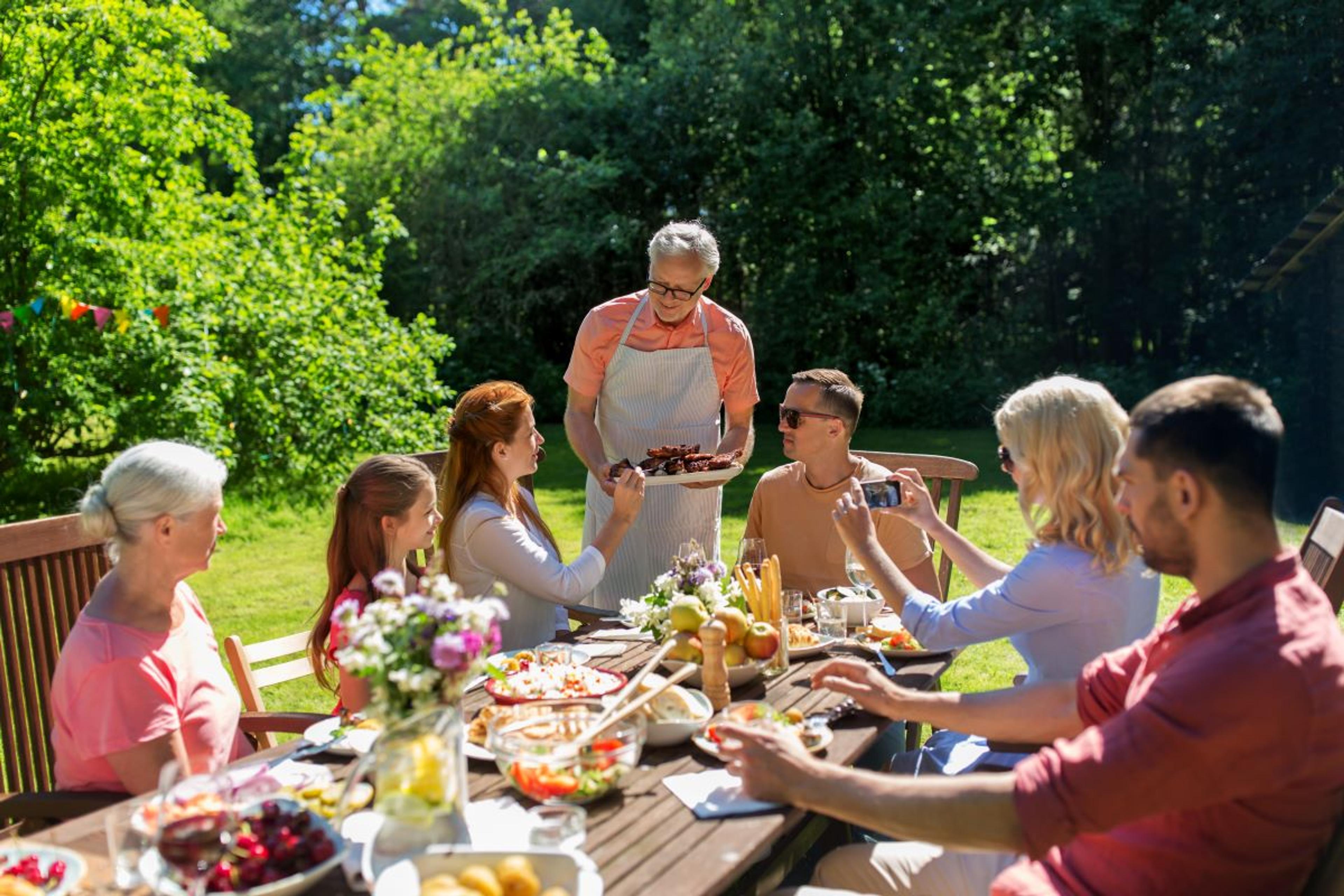 Friends smiling as they pass dishes to each other at a table outside in the yard.