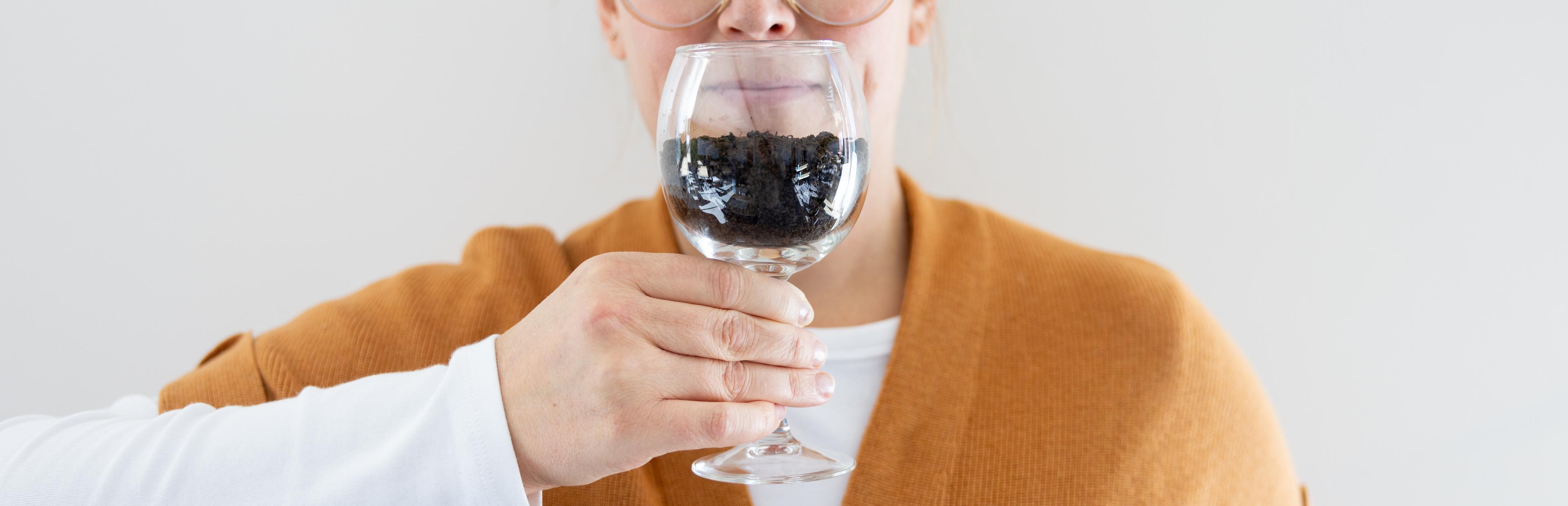 A woman sniffs a wine glass that is half full of soil.