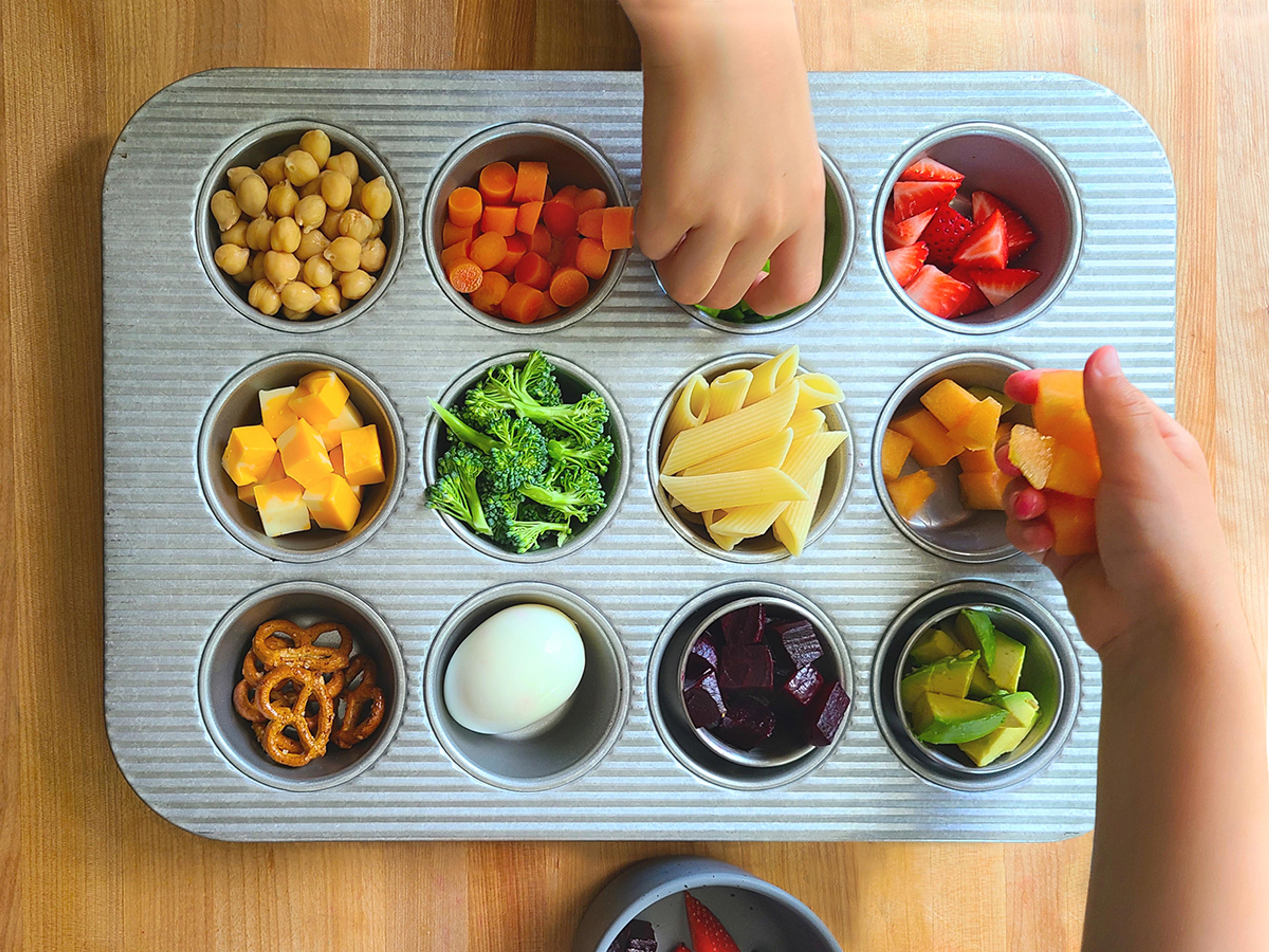  Two children’s hands fill each well of a muffin tin with a different colorful food.
