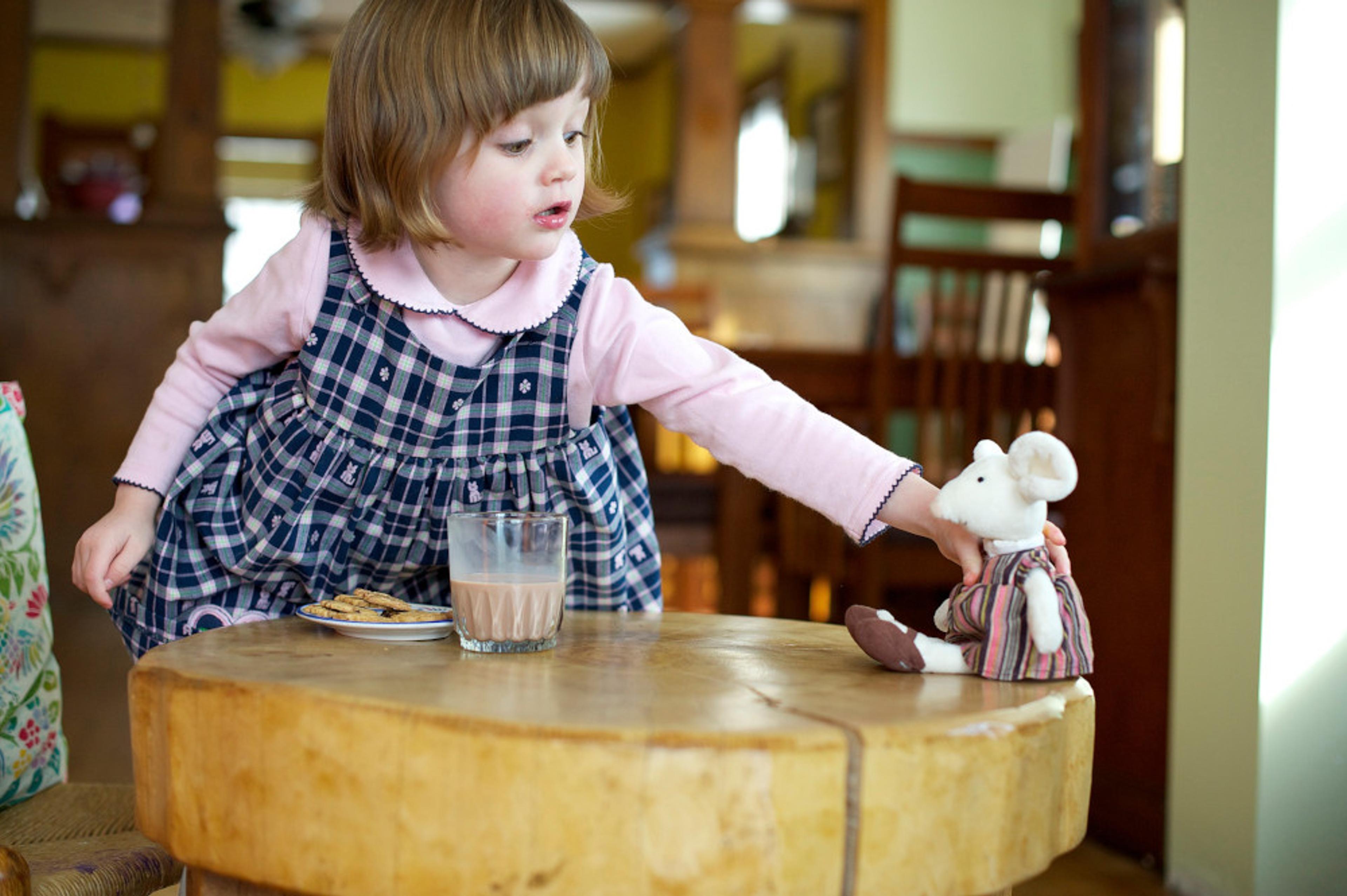 A little girl playing with a stuffed animal while having a glass of chocolate milk.