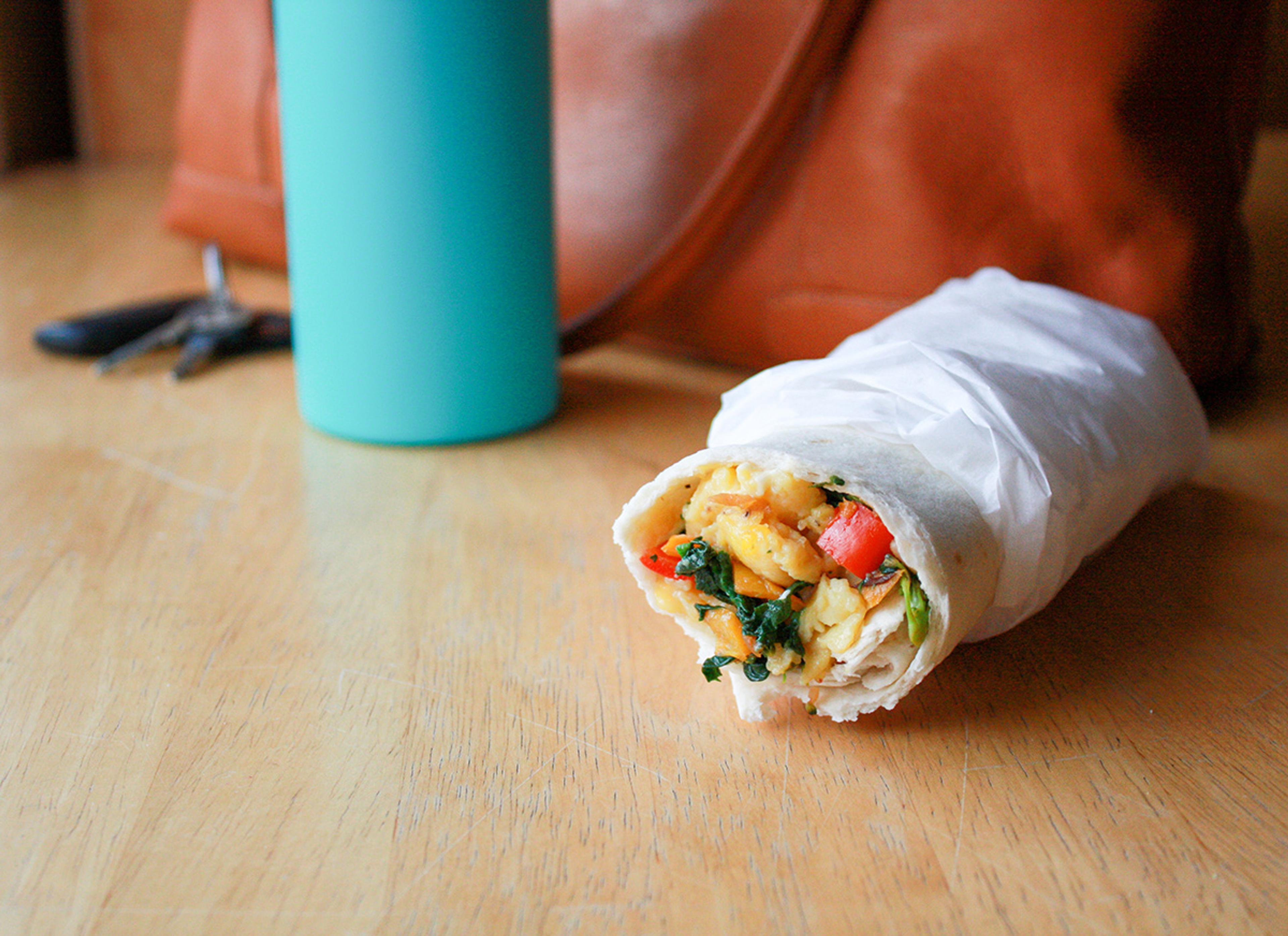 A wrapped breakfast burrito with the end open to show the filling sits on a table in front of a woman’s leather purse, water bottle and car keys.
