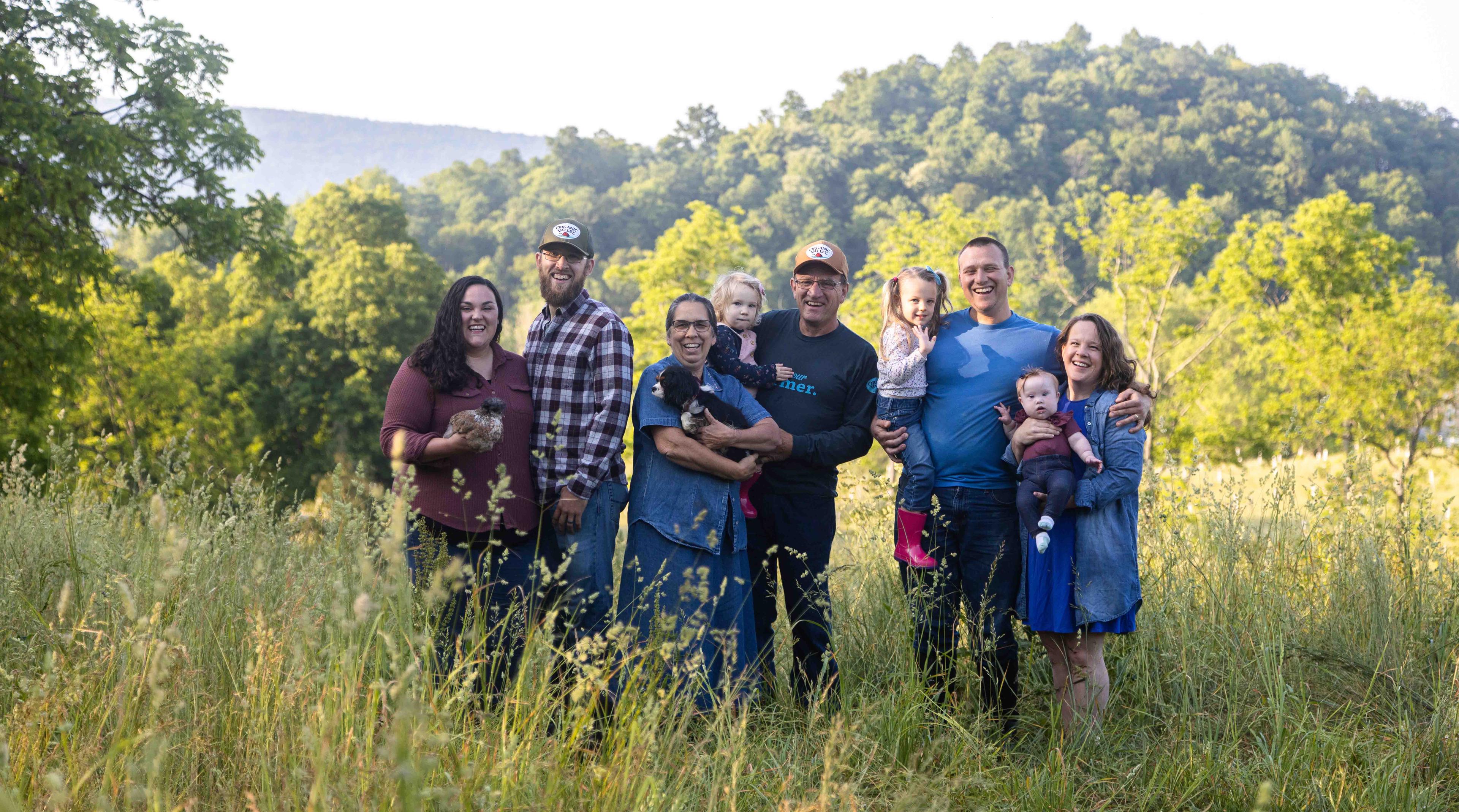 Family of farmers standing in field holding children and pets with rolling hills in the background. 