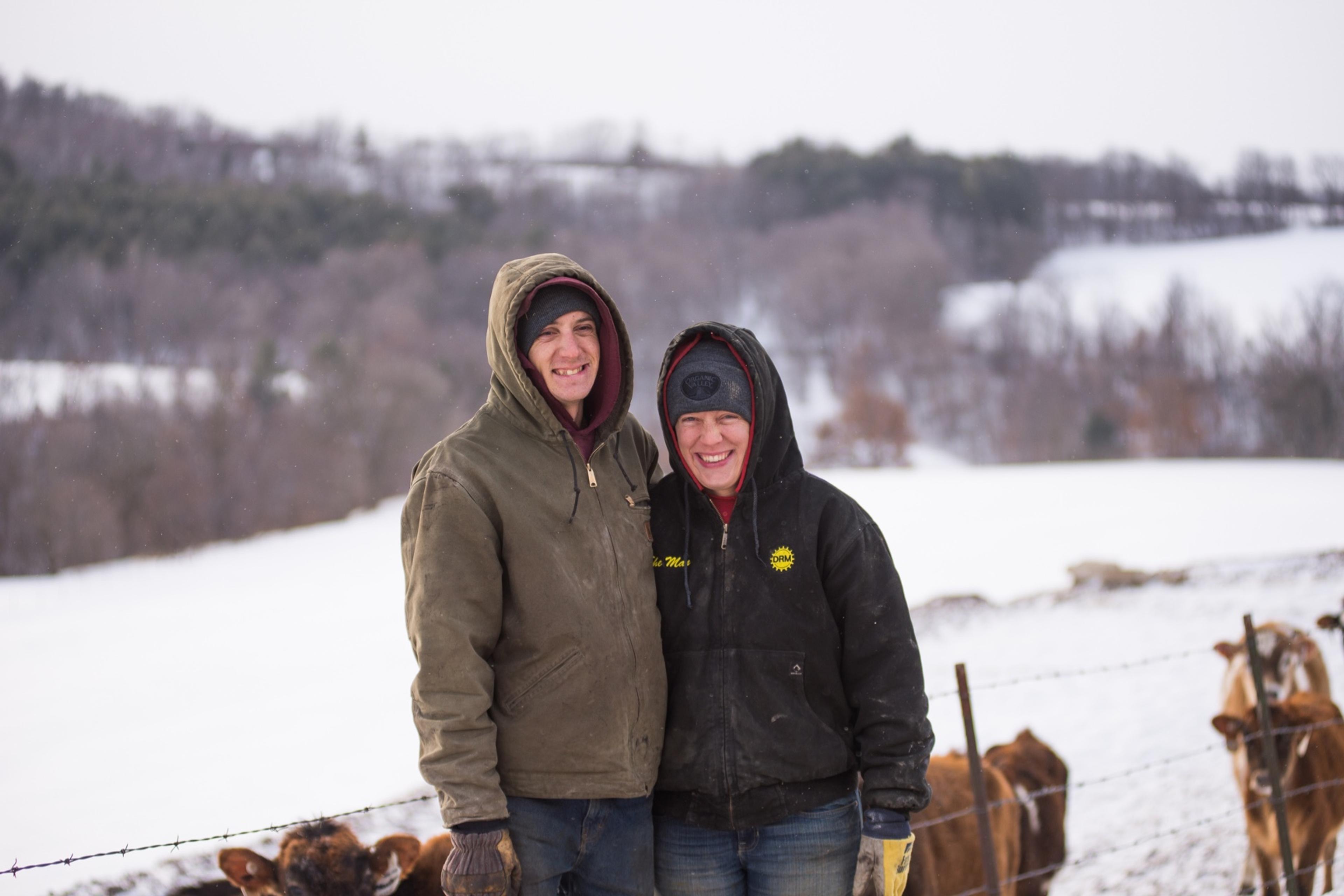 Bundled up in coats, Amy and Marques Koenig smile and look into the camera while standing in front of a snowy pasture with cows.