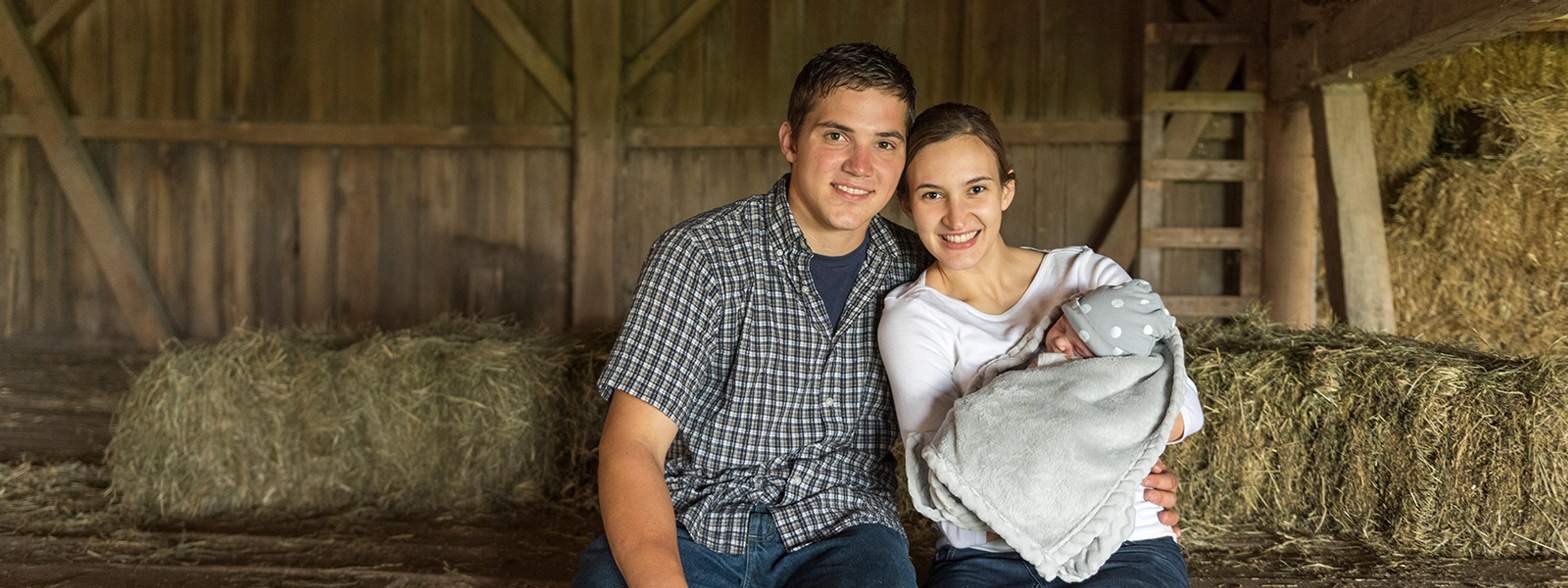 Young farmers posing in a barn with their newborn child.