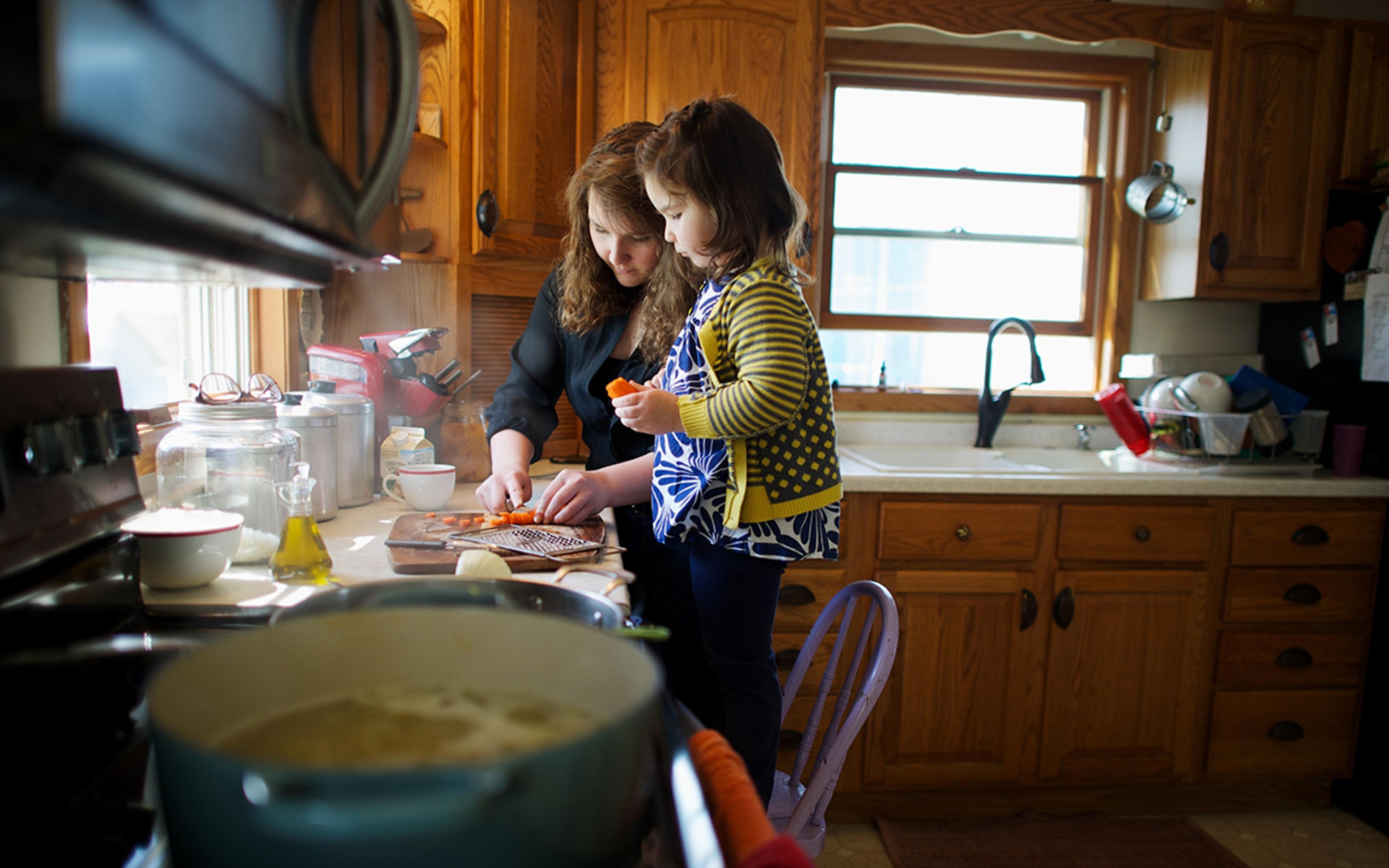 A girl stands on a chair at the kitchen counter and watches her sister chop carrots. A pot of soup simmers on the stove.