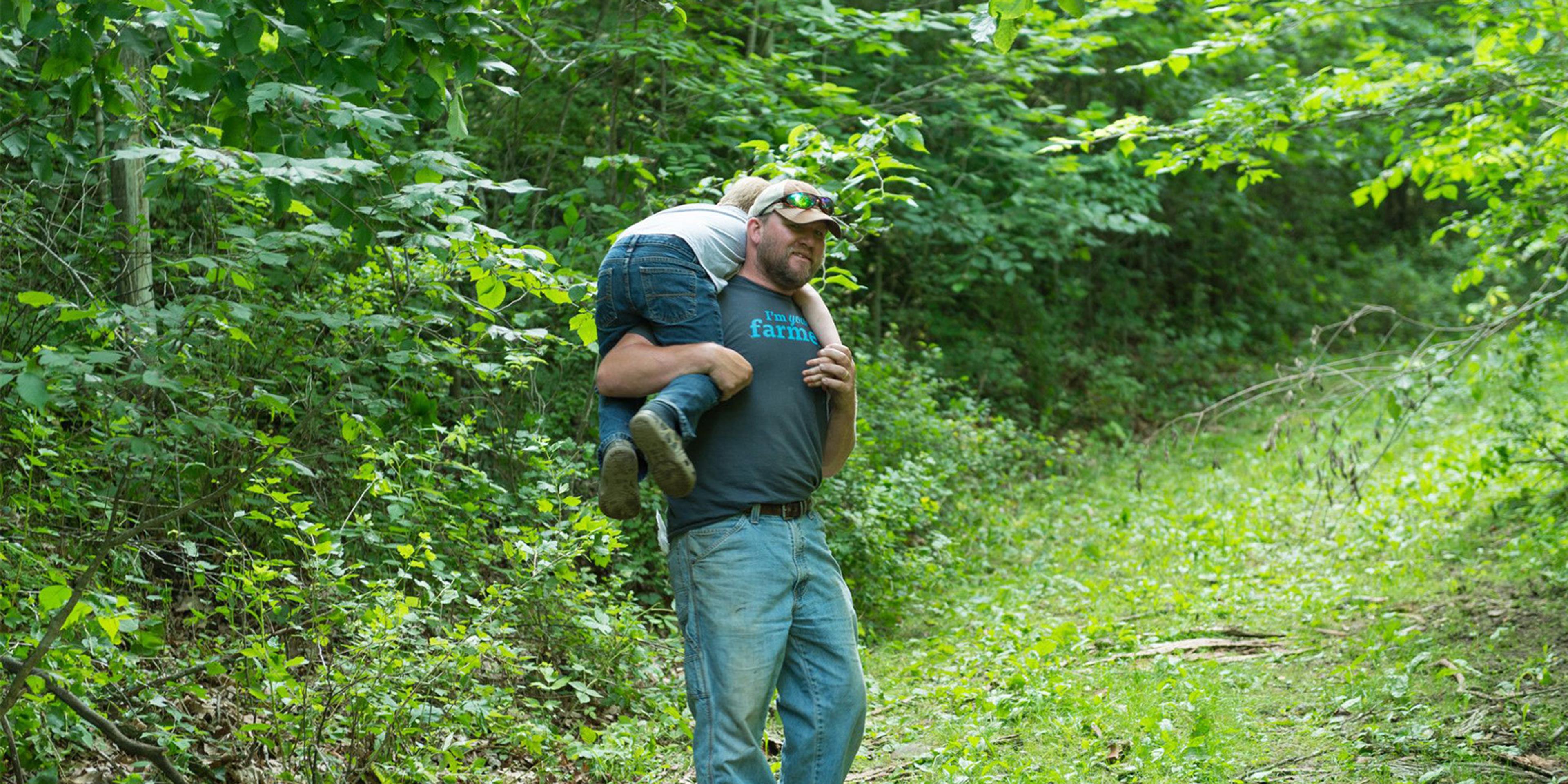 Farmer Tucker Gretebeck carries his son in a wooded area on his Wisconsin farm.