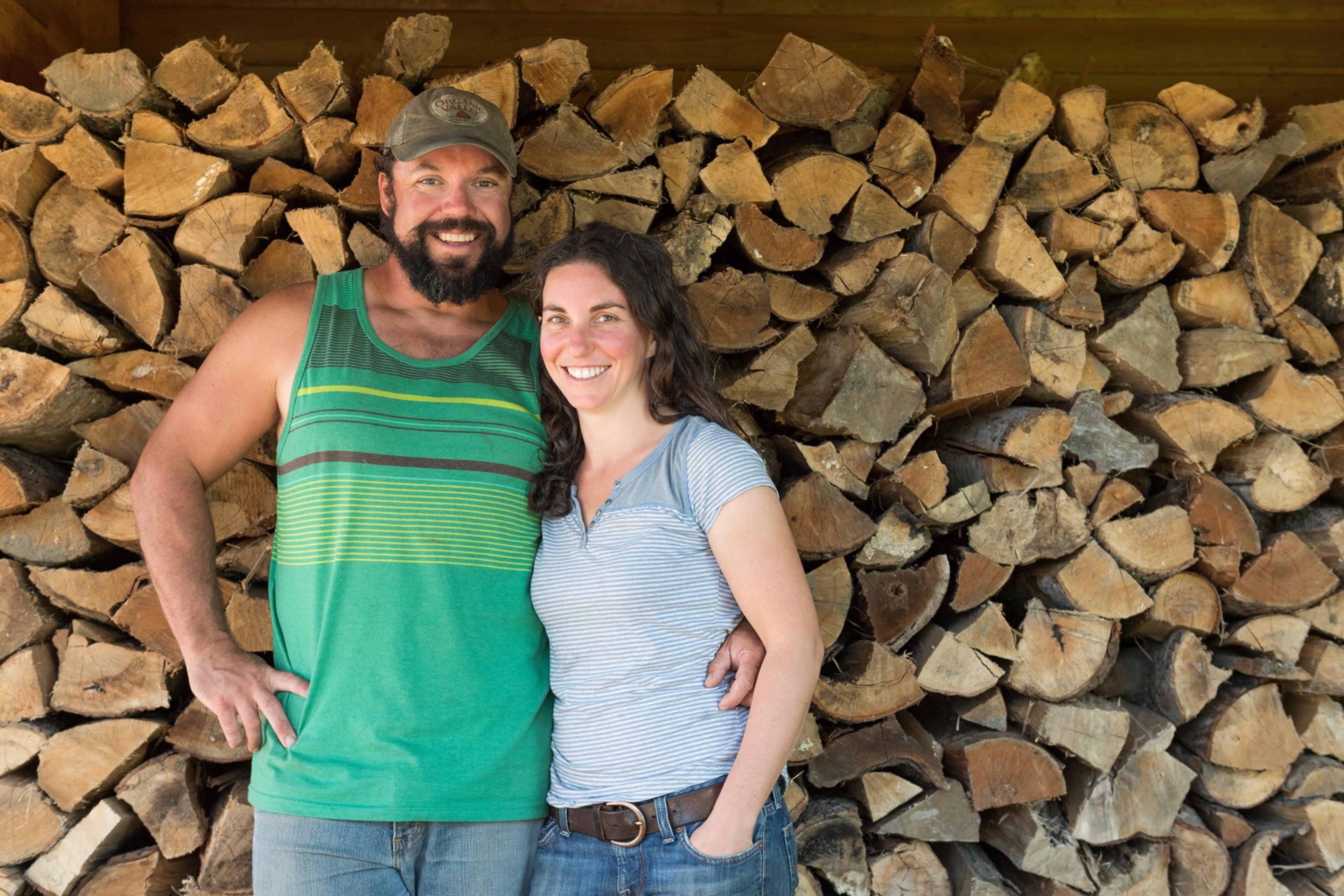 Tyler and Melanie smile at the camera while standing in front of a large stack of firewood.