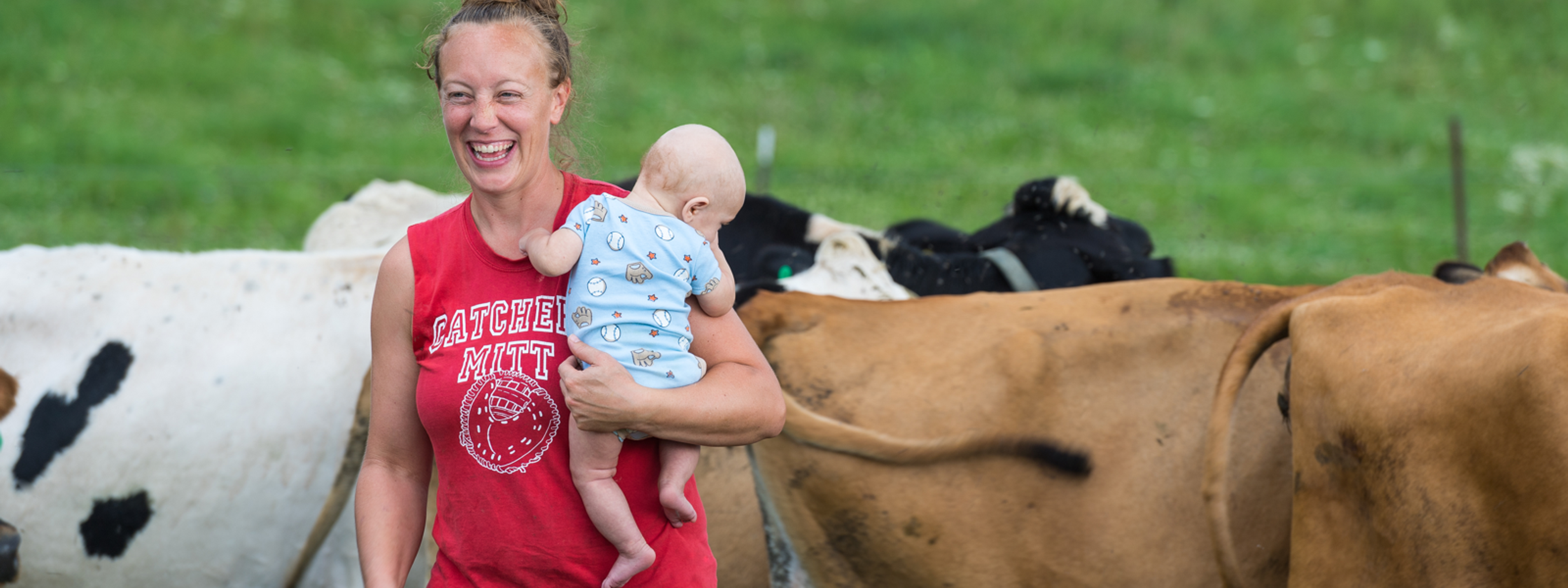 Amy Koenig has a big smile while carrying her baby through a pasture with cows behind her.