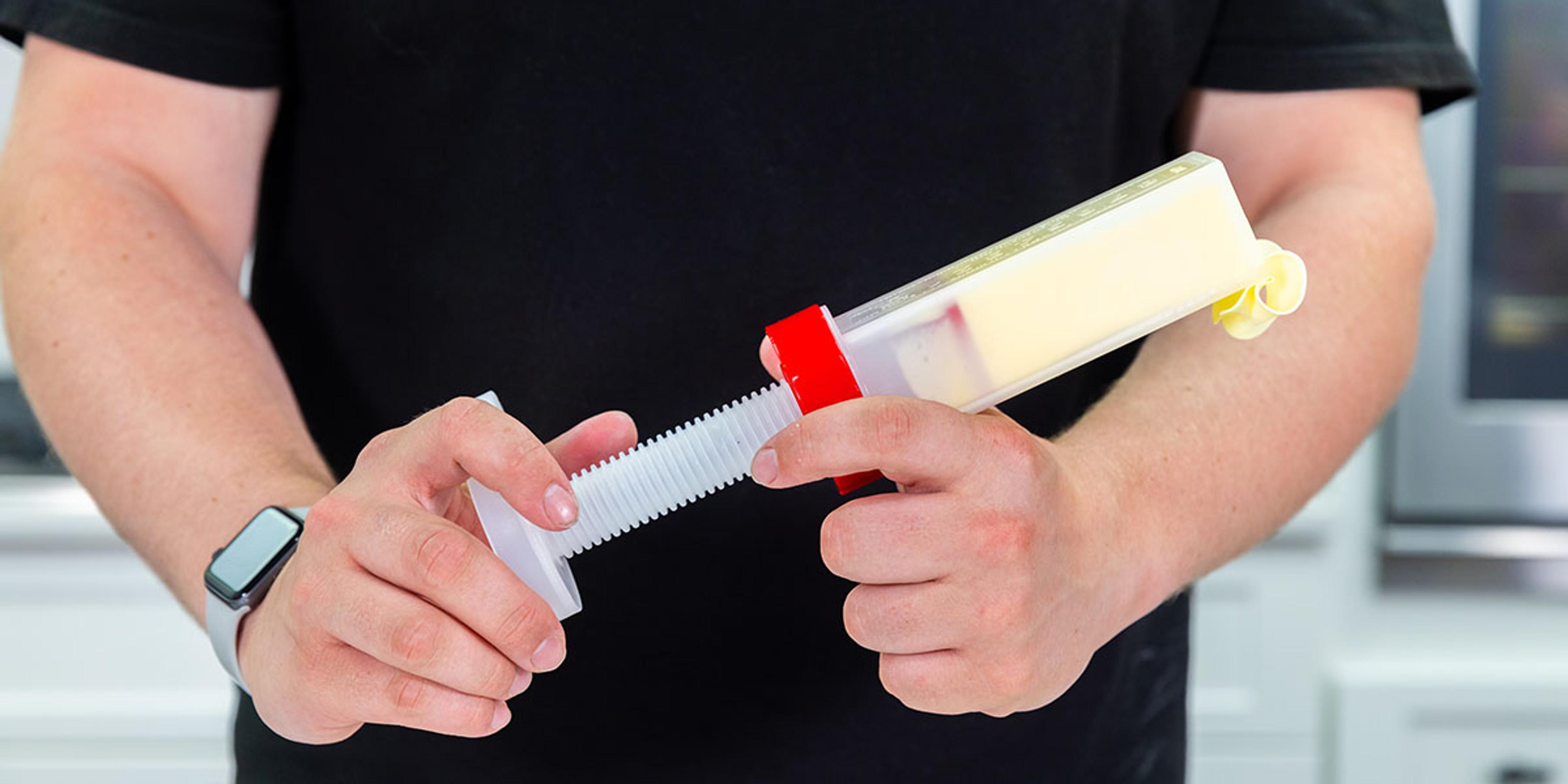 A man holds a plastic butter dispenser with a twist handle as butter oozes out the top.