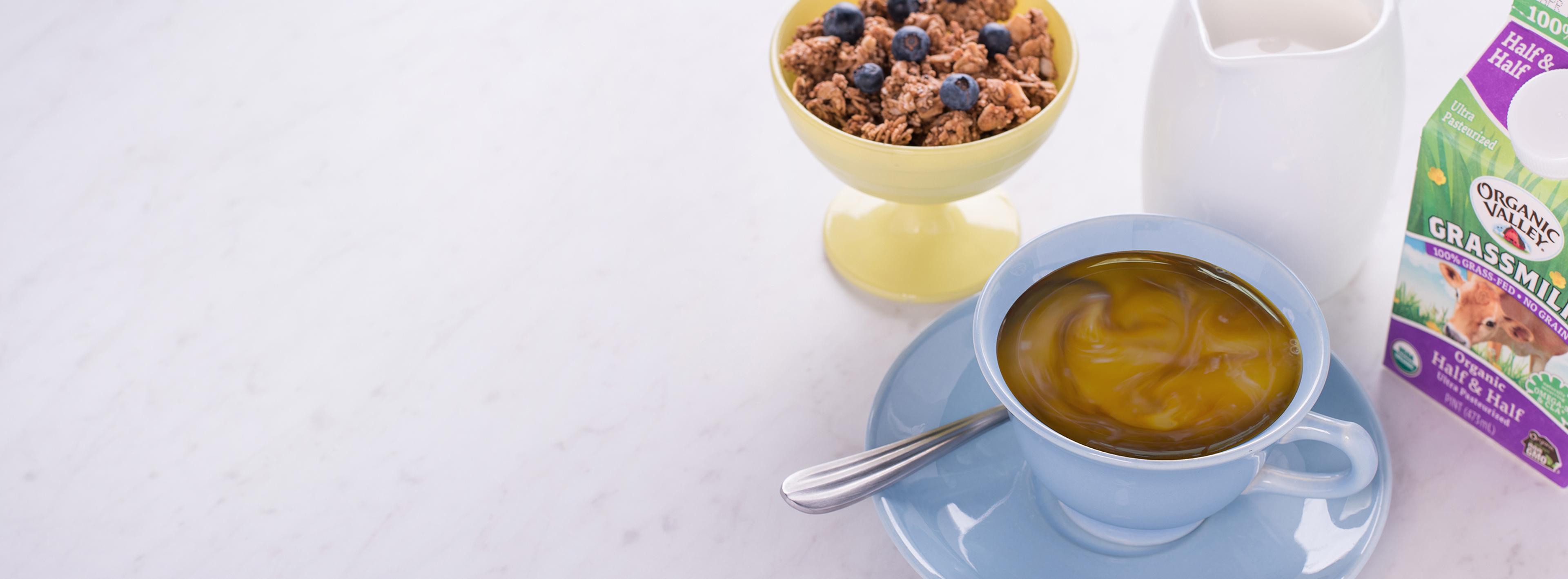 A cup of coffee with creamer in it next to a bowl of granola. 