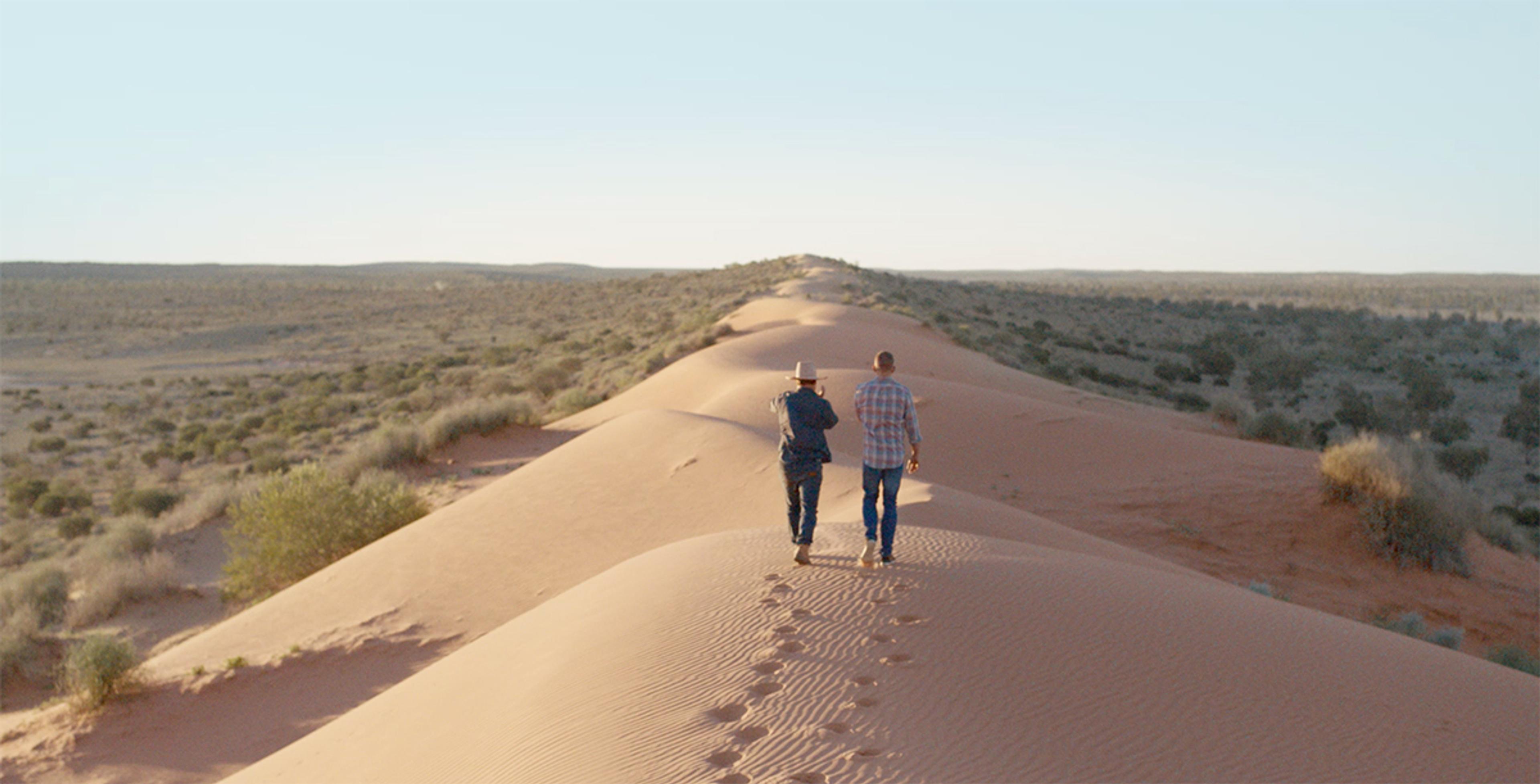 Two men walk along a sand dune rising up from the Australian grasslands.