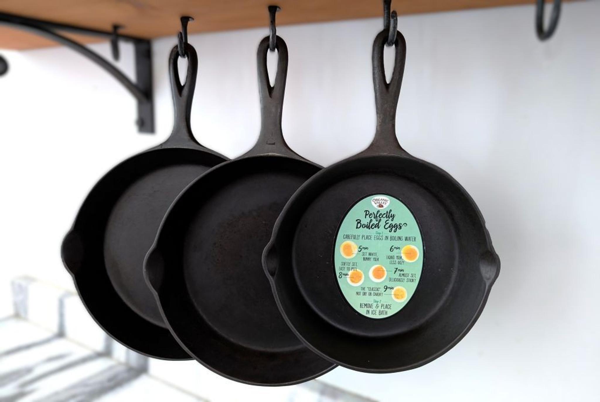 Three cast iron frying pans hang on a floating shelf; one pan has a guide to cooking hard boiled eggs magnet stuck it.