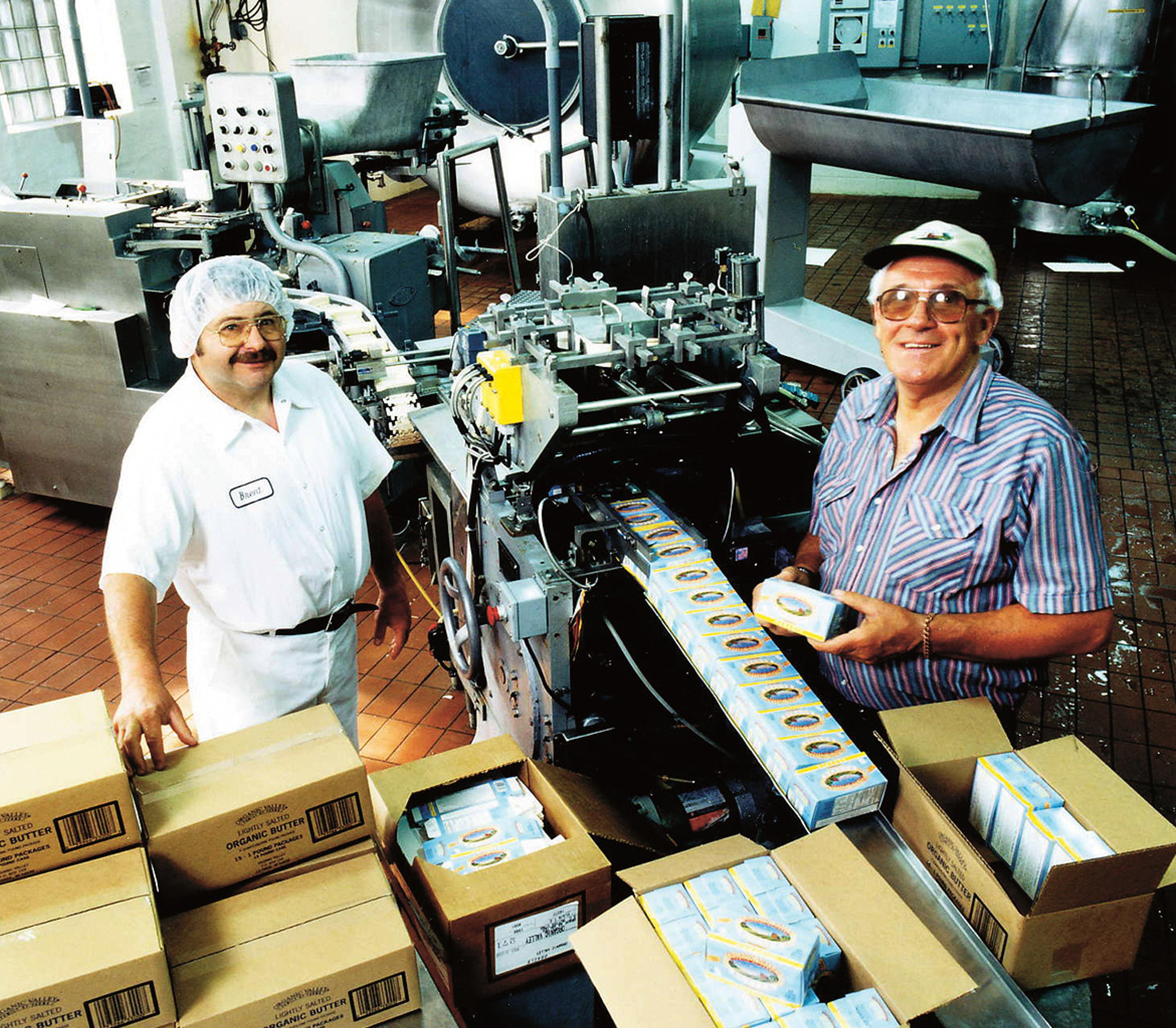 Two men stand in the creamery next to a butter packaging machine and many partially filled shipping boxes.