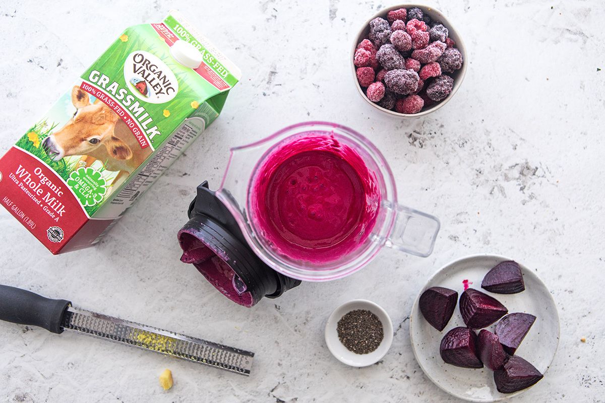 Overhead view of a berry beet smoothie in a blender surrounded by an Organic Valley Grassmilk carton, frozen berries, quartered beets, and fresh grated ginger.. 