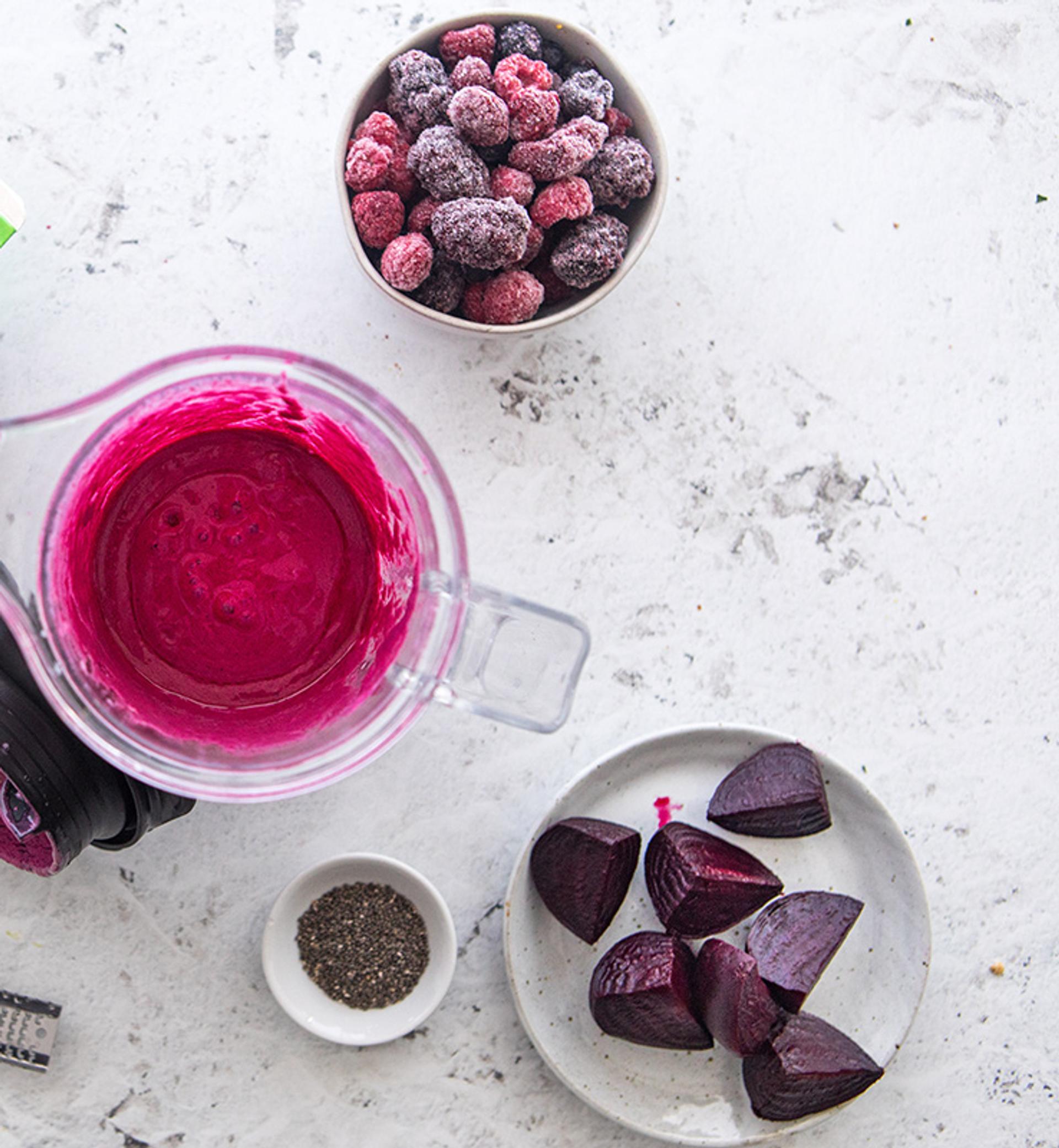 Overhead view of a berry beet smoothie in a blender surrounded by an Organic Valley Grassmilk carton, frozen berries, quartered beets, and fresh grated ginger.. 