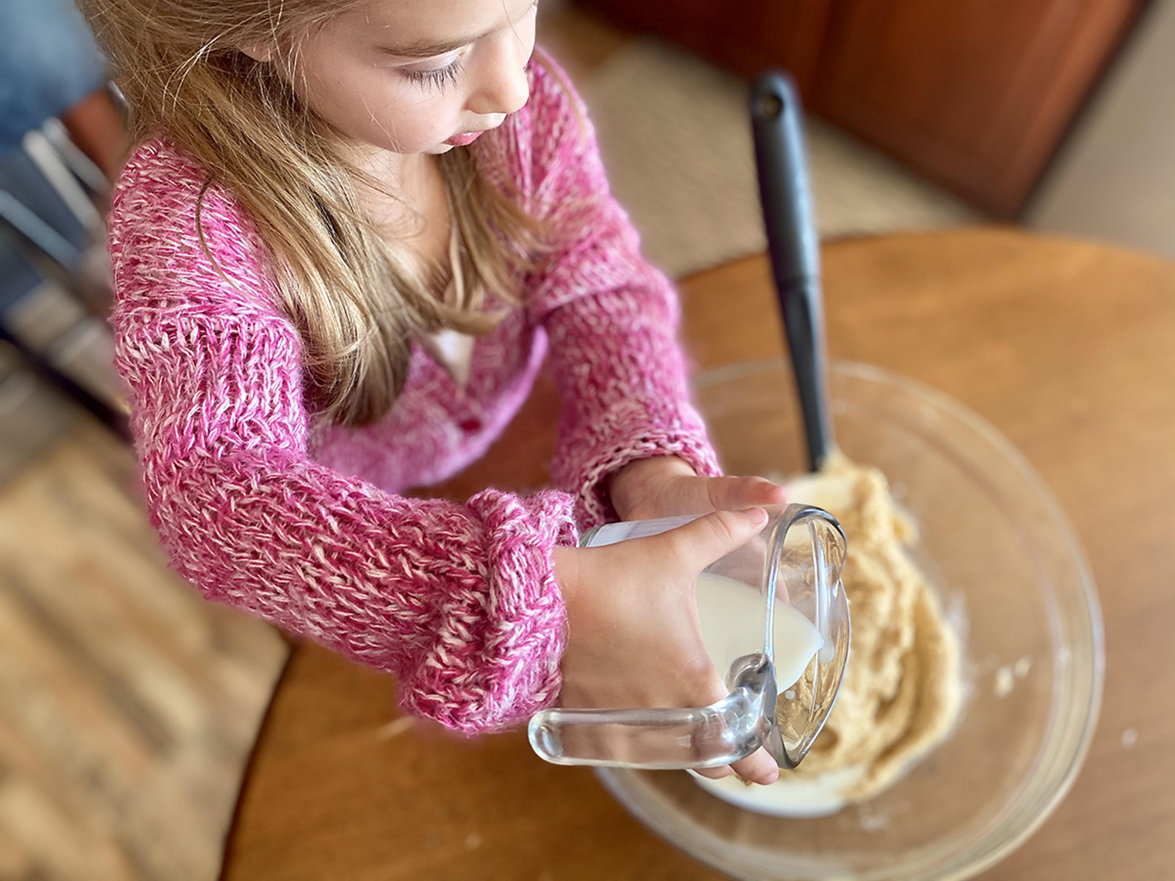 Overhead view of the hands of a little girl in a pink sweater pouring milk from a measuring cup into a mixing bowl. 