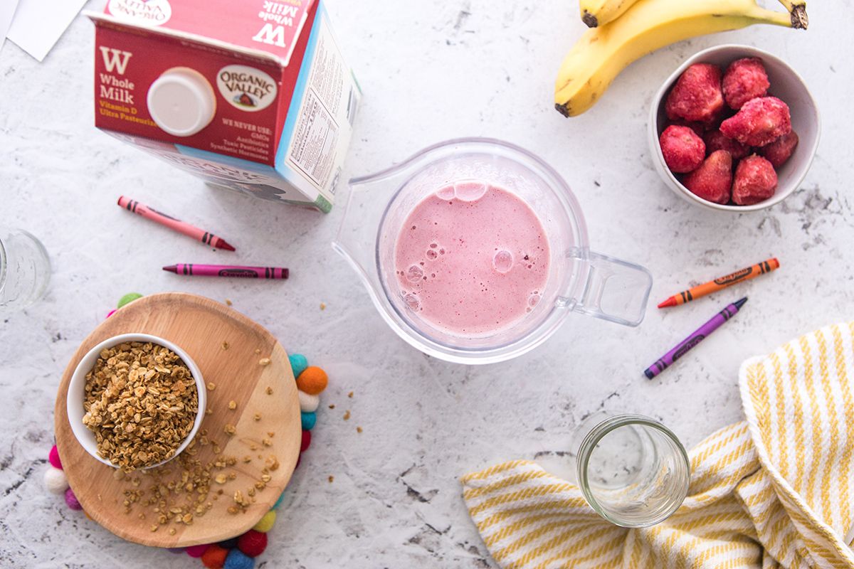 Overhead view of a smoothie in a cup surrounded by an Organic Valley milk carton, frozen strawberries, granola, and crayons for decor.