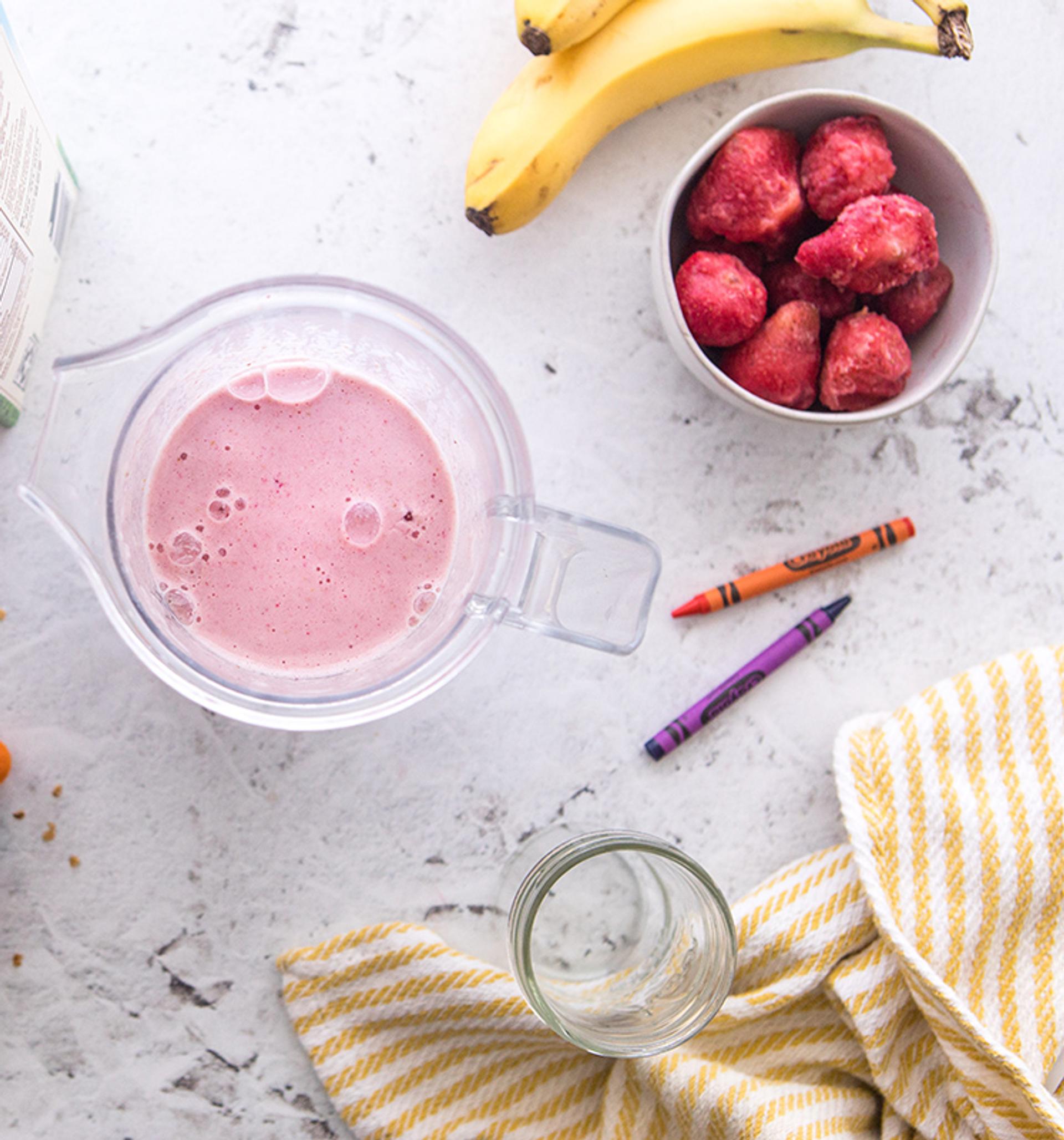 Overhead view of a smoothie in a cup surrounded by an Organic Valley milk carton, frozen strawberries, granola, and crayons for decor.