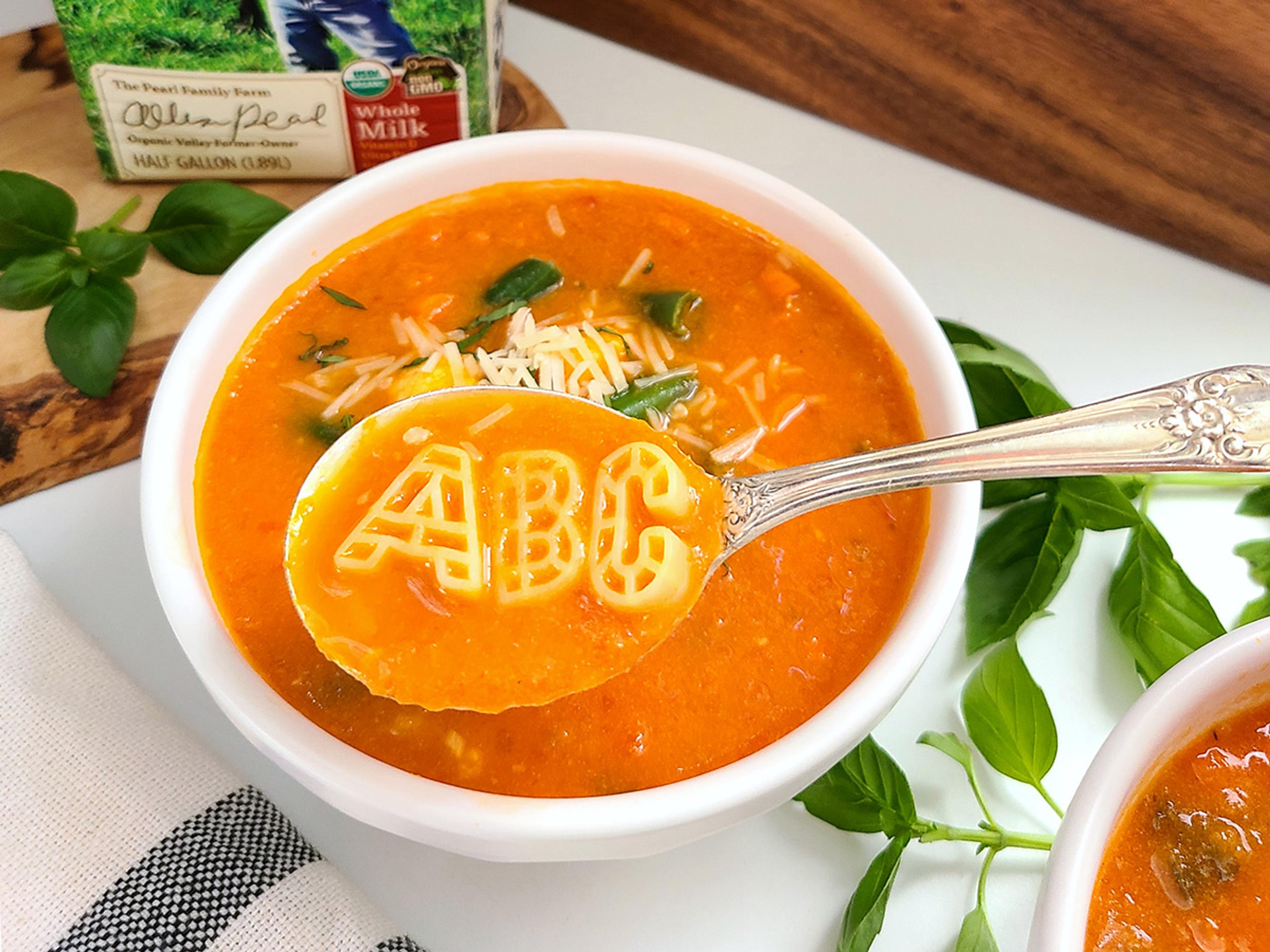 A spoon holds A, B, C pasta over a bowl of tomato-based soup sprinkled with shredded cheese.
