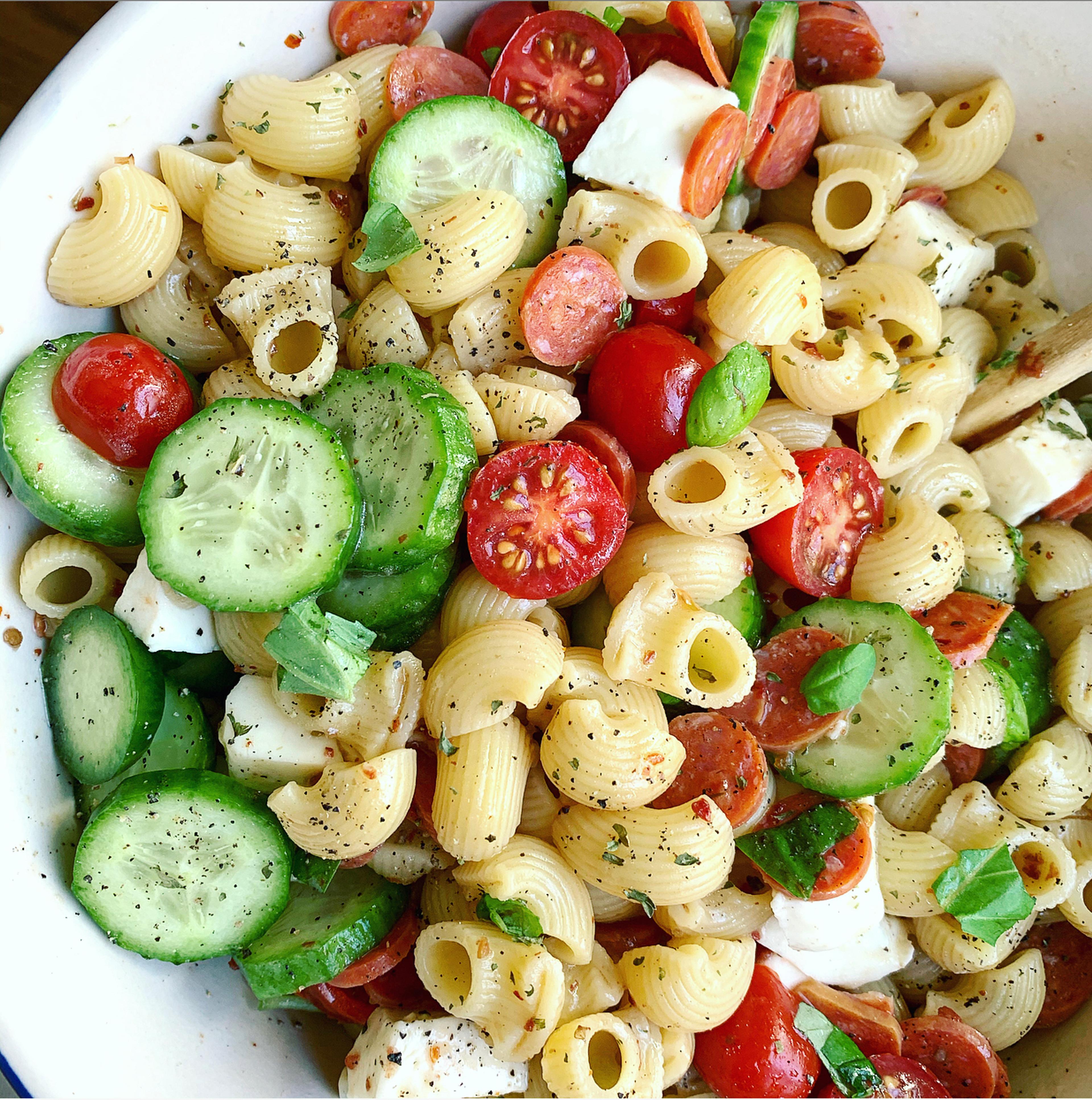 A colorful pasta salad with elbow pasta, cherry tomatoes, sliced cucumbers, mini pepperonis, and cubed cheese sprinkled with pepper.