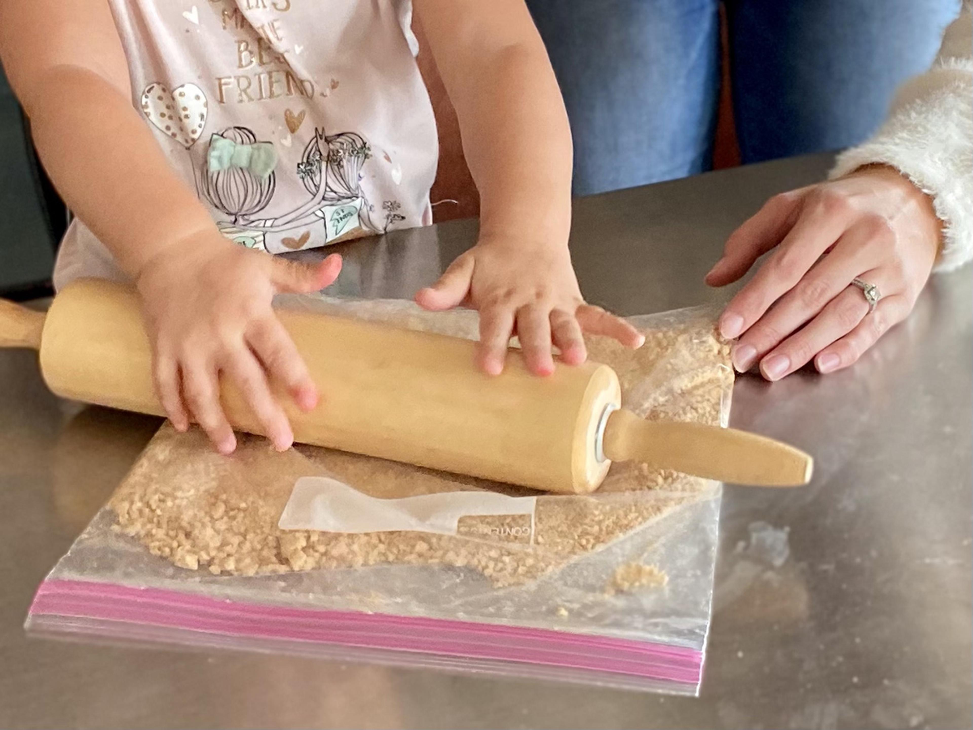 View of a little girl's hands using a rolling pin to smash crackers in a plastic bag.