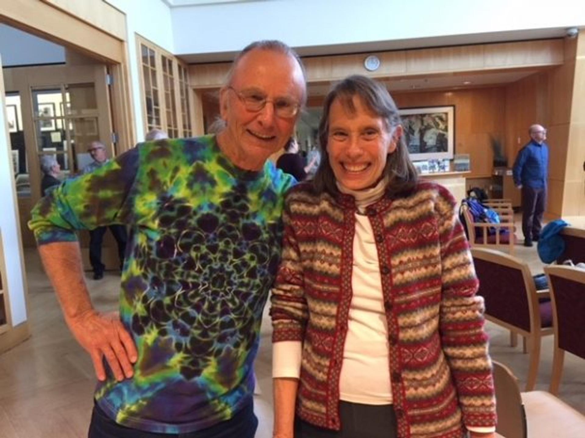 Harry MacCormack wears a tie dyed shirt and smiles with Lynn Coody in what looks like the meeting room of a library.