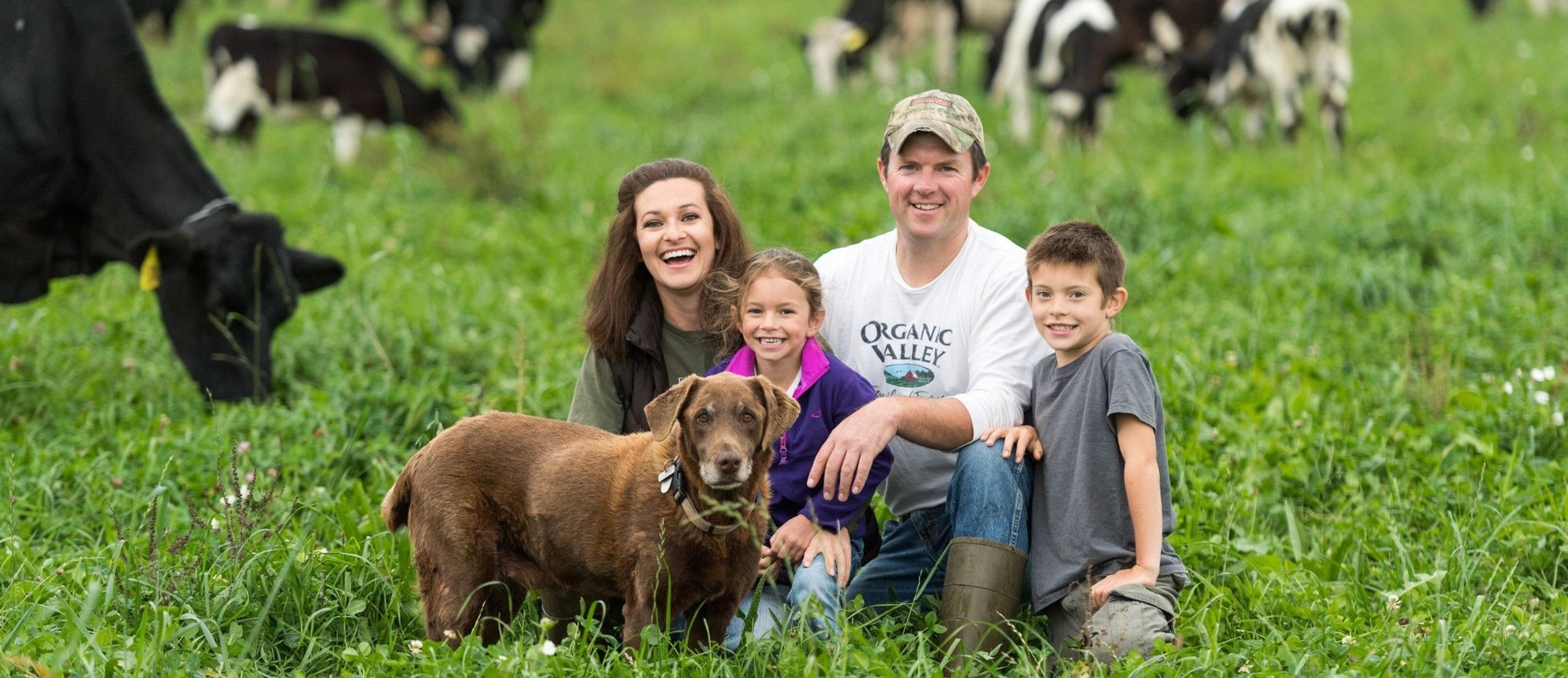 Four members of the Huftalen family sit on pasture with their dog and cows on their organic dairy farm in New York.