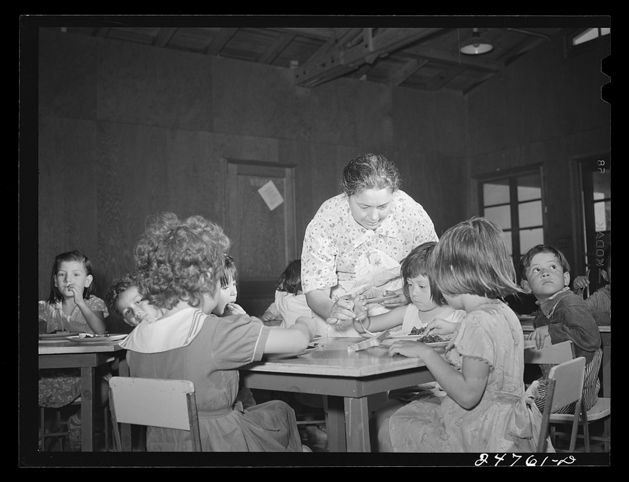 A black and white photo of children in old fashioned clothing sitting at tables being served food by a woman. (Source: Library of Congress)