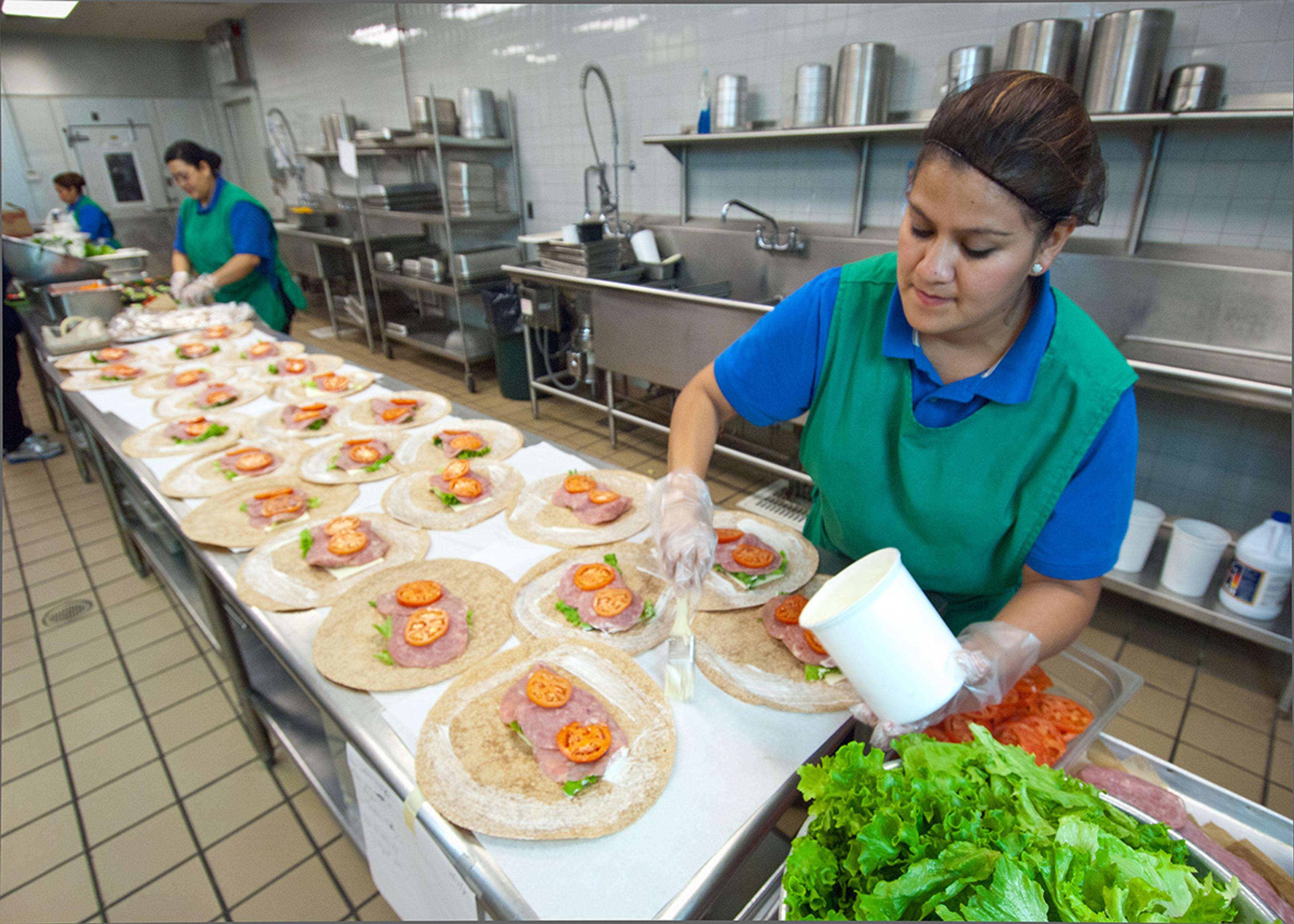 A school lunchroom cook spreads mayo onto meat and veggie wraps that she's preparing in the school kitchen.