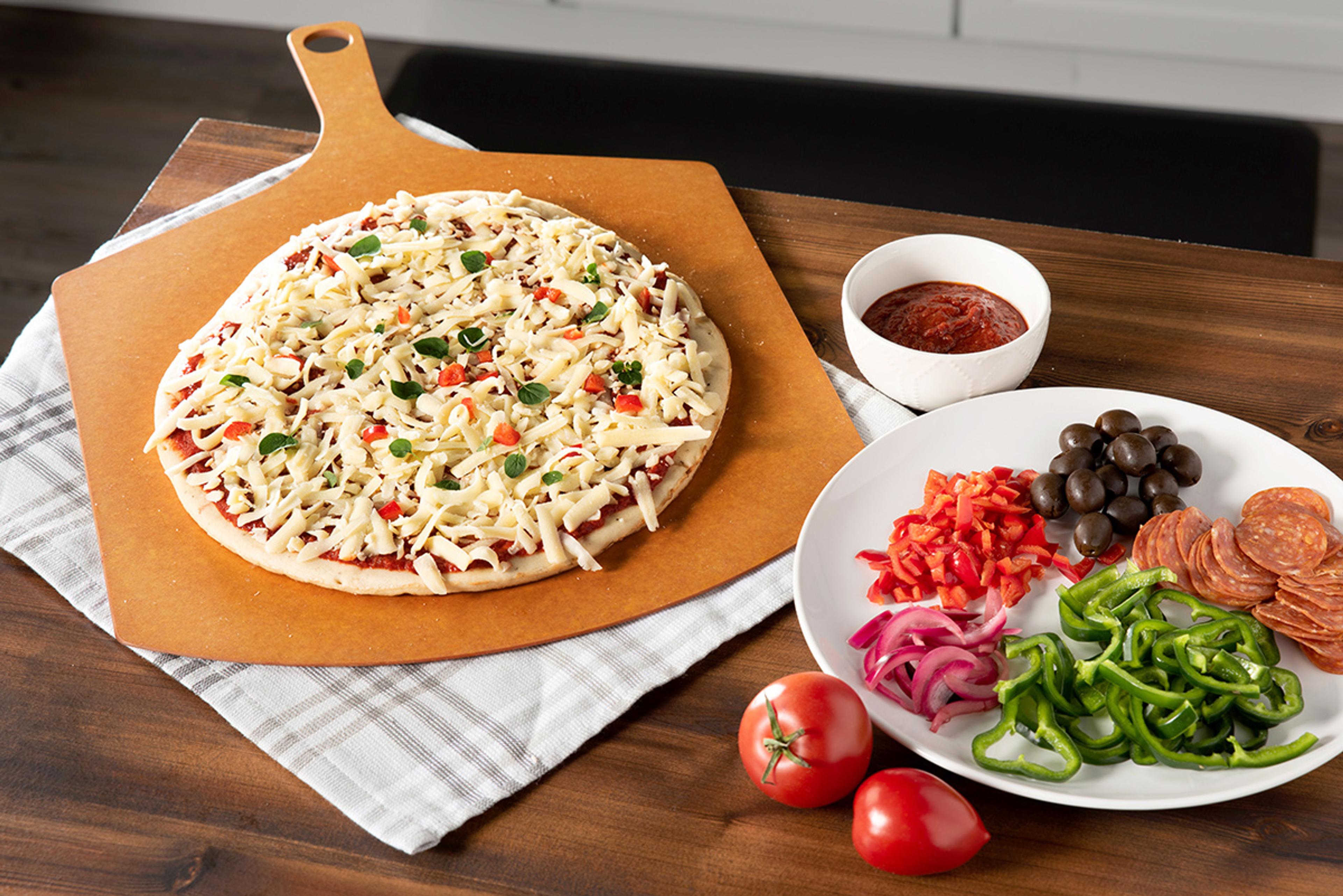 An unbaked pizza sits on a pizza peel with a plate of toppings sitting next to it.