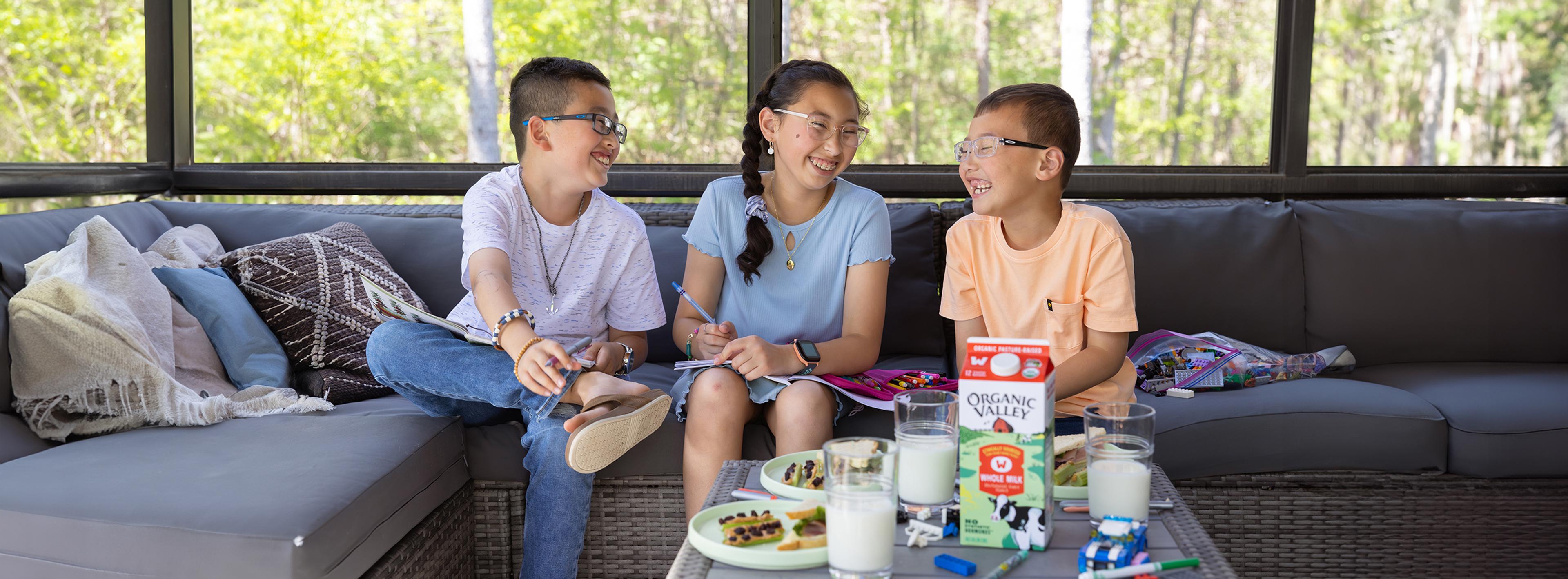 Three children on couch smiling while drinking milk and eating snacks.