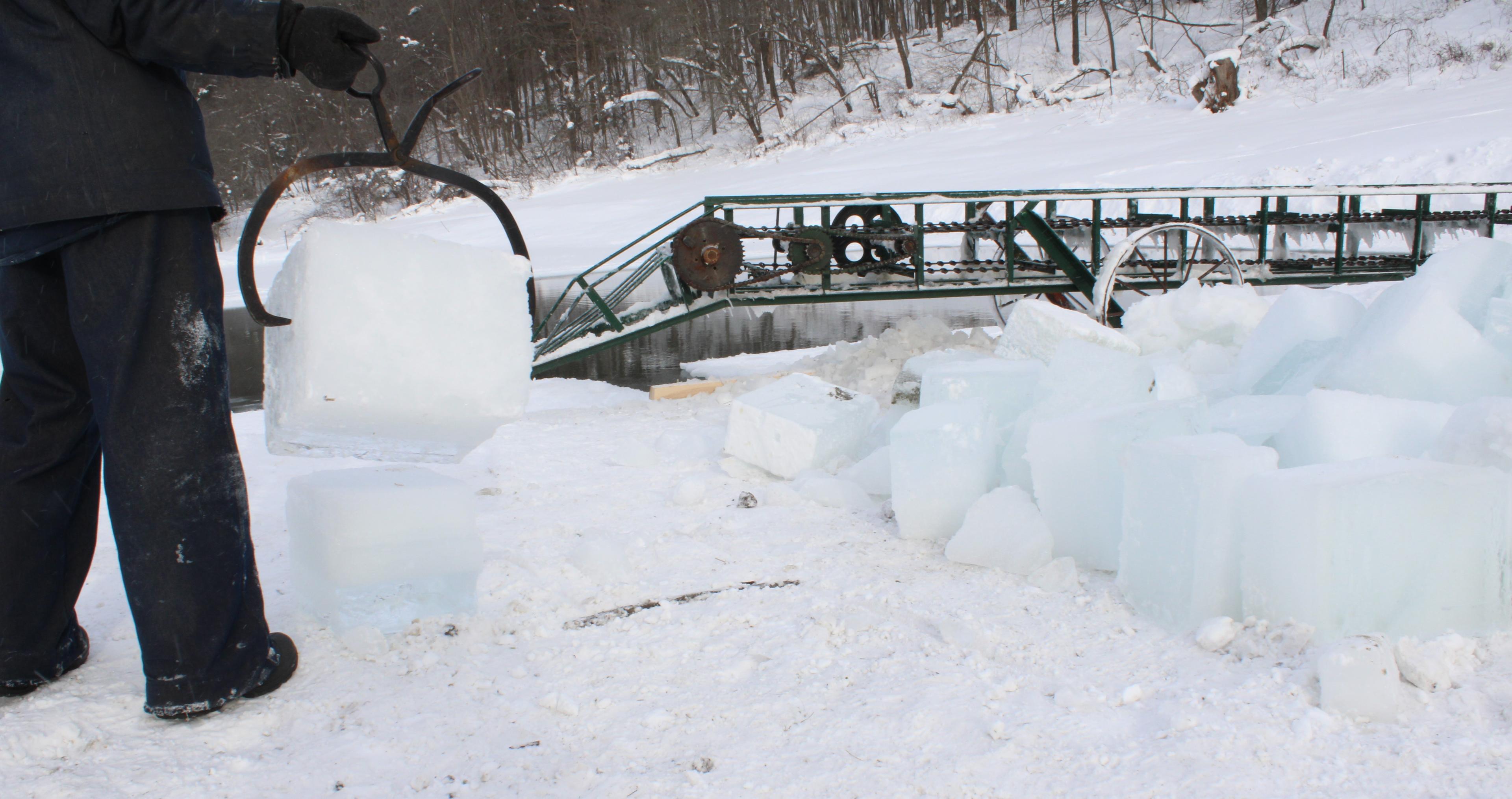 A man holds an ice block in tongs and blocks of ice are piled in front of him.