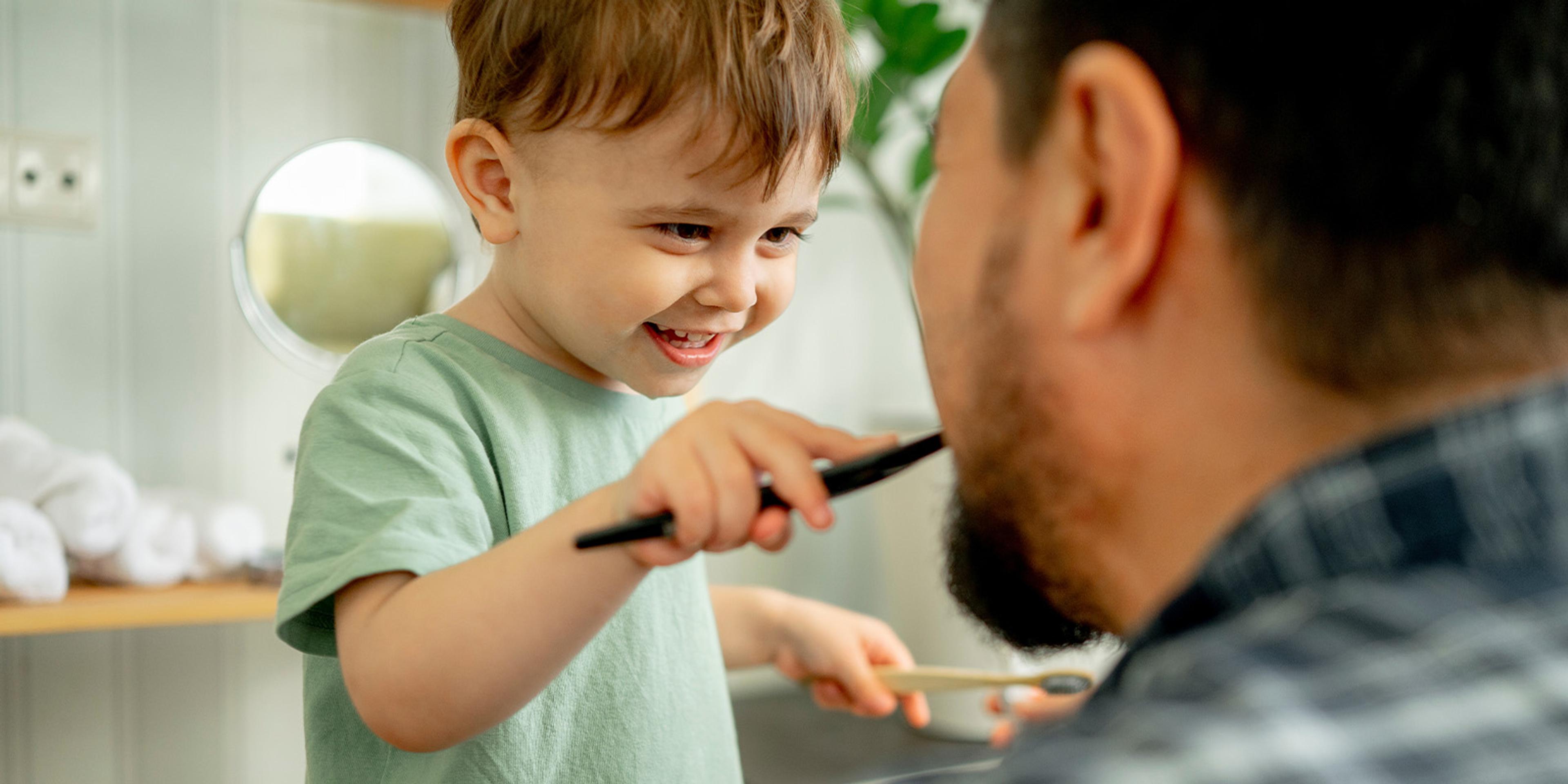 A toddler brushes a dad’s teeth and holds two toothbrushes.