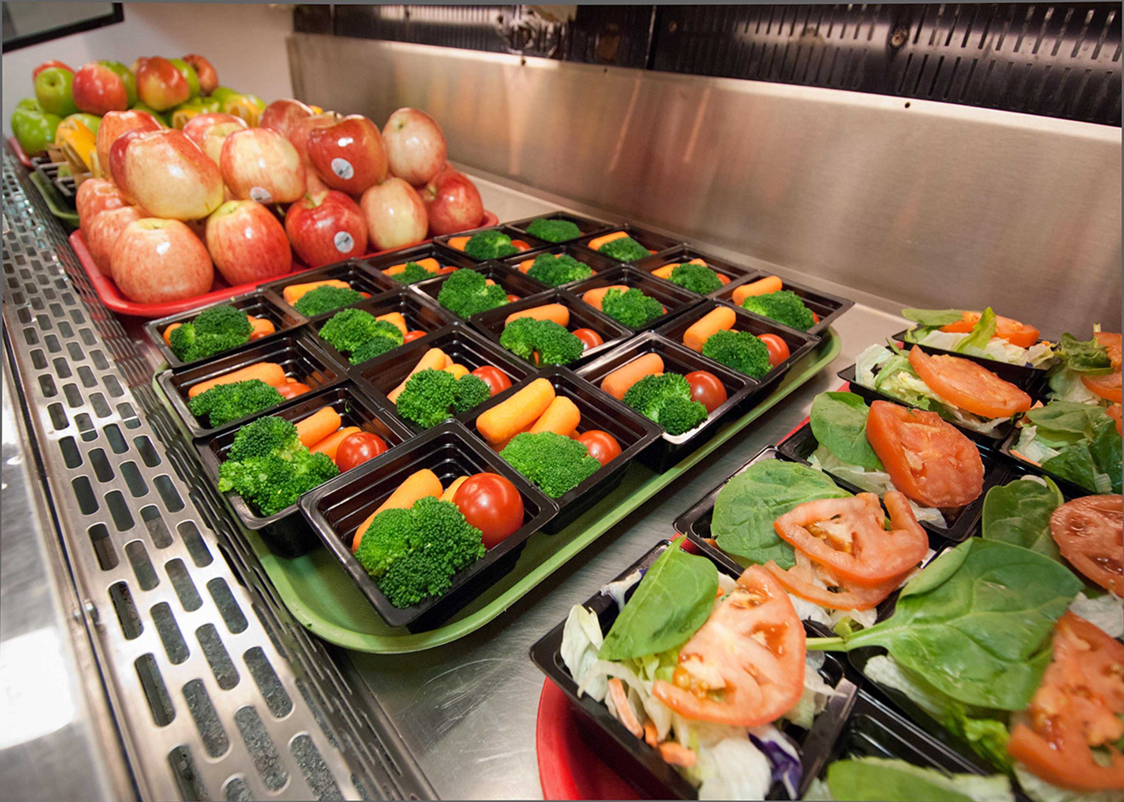 Prepared containers of colorful fresh fruits and vegetables lined up on a school lunch bar.