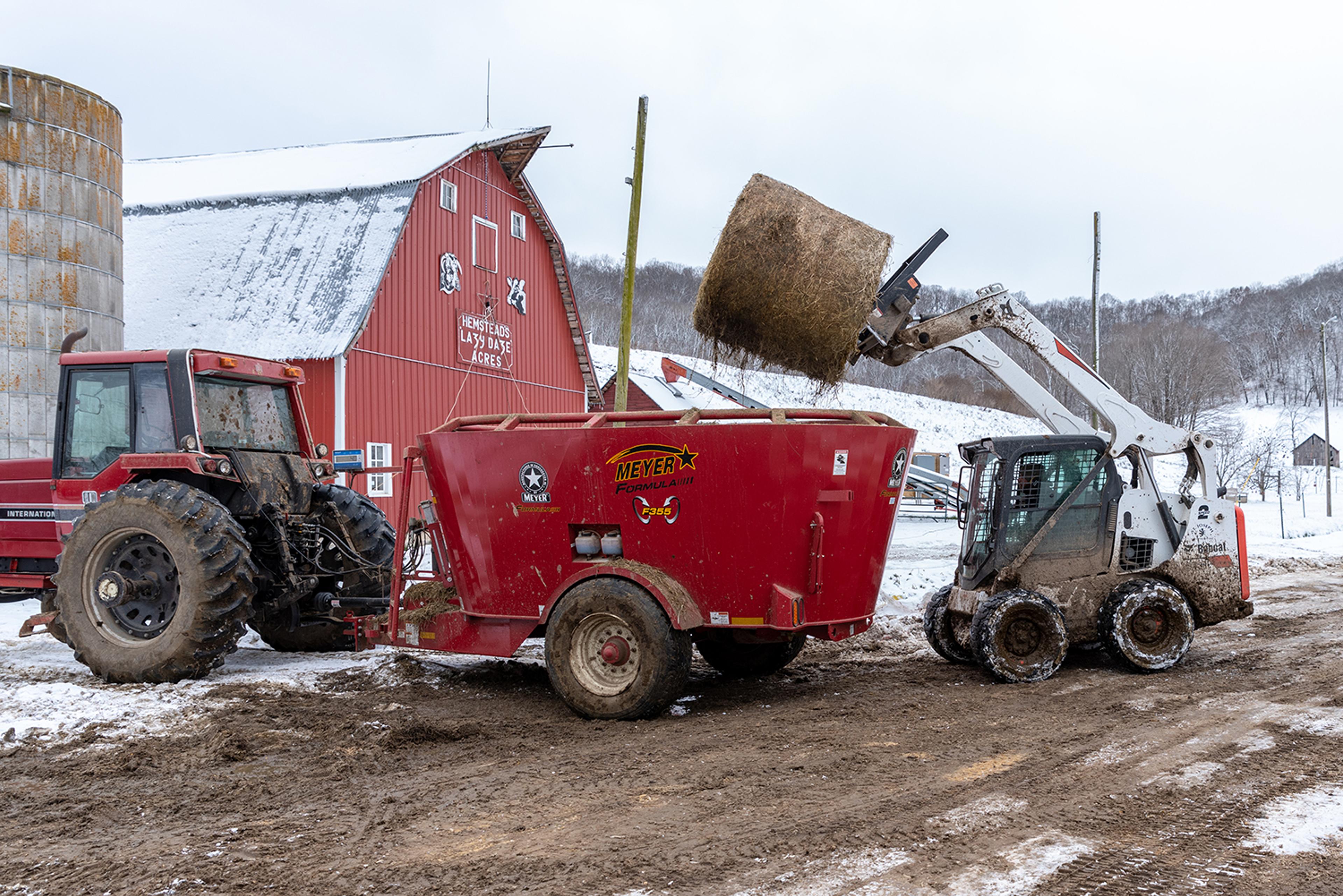 A farmer uses a Bobcat to load a round bale of hay into a trailer pulled by a big tractor. 