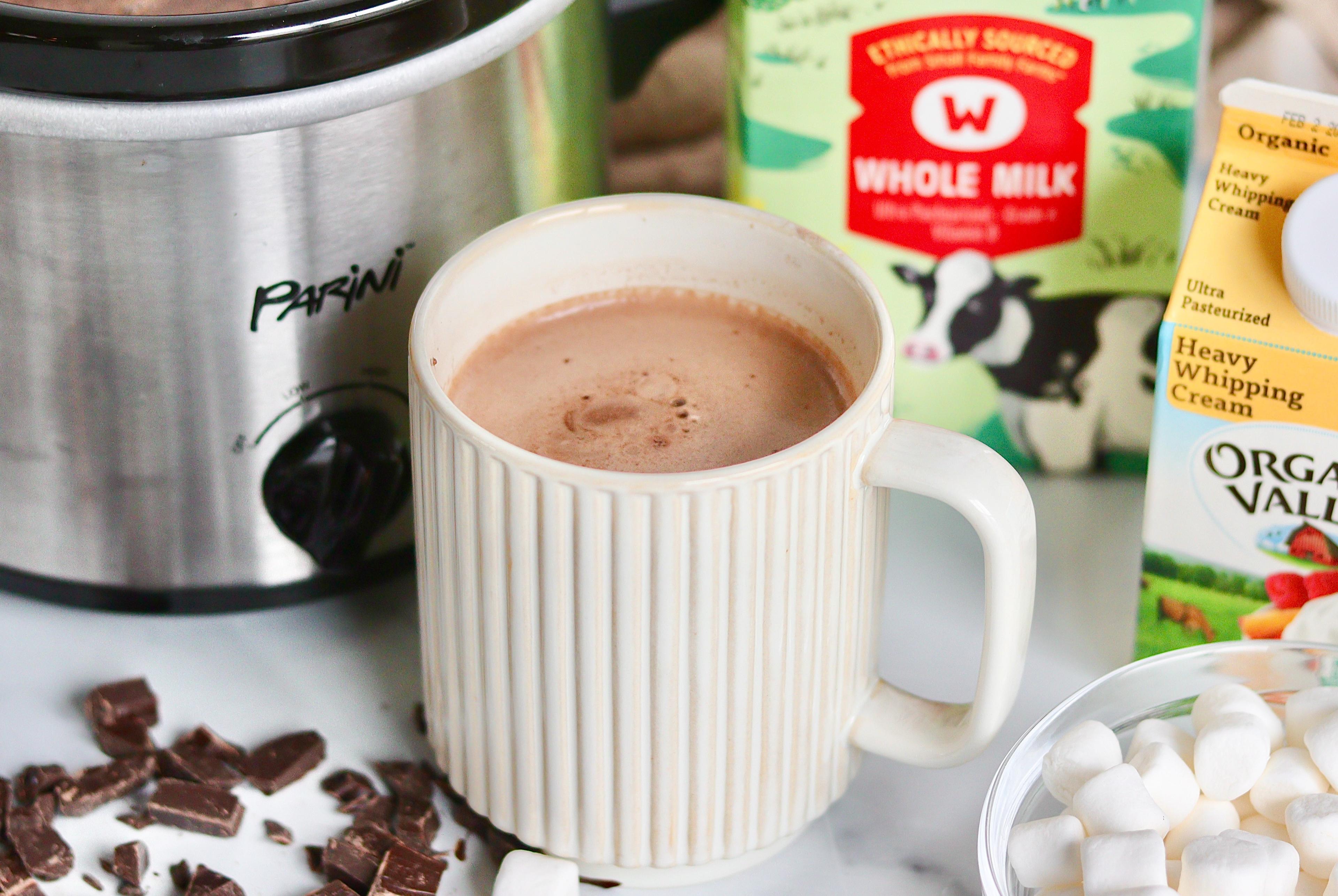 A mug of hot chocolate next to a slow cooker, chocolate shavings, marshmallows and organic dairy products used to make hot chocolate.